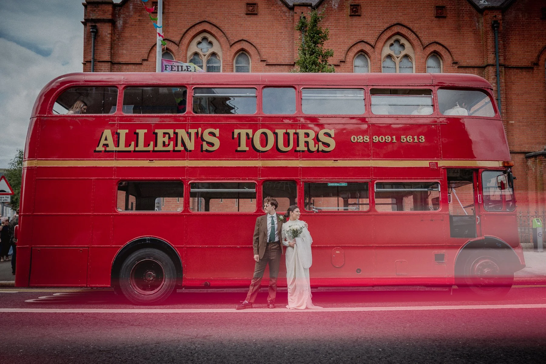 A red double-decker tour bus with the sign 'ALLEN'S TOURS' and a phone number. Two people in wedding attire, a man and a woman, stand in front of the bus, holding hands. The woman is holding flowers. The background features a brick church with arched