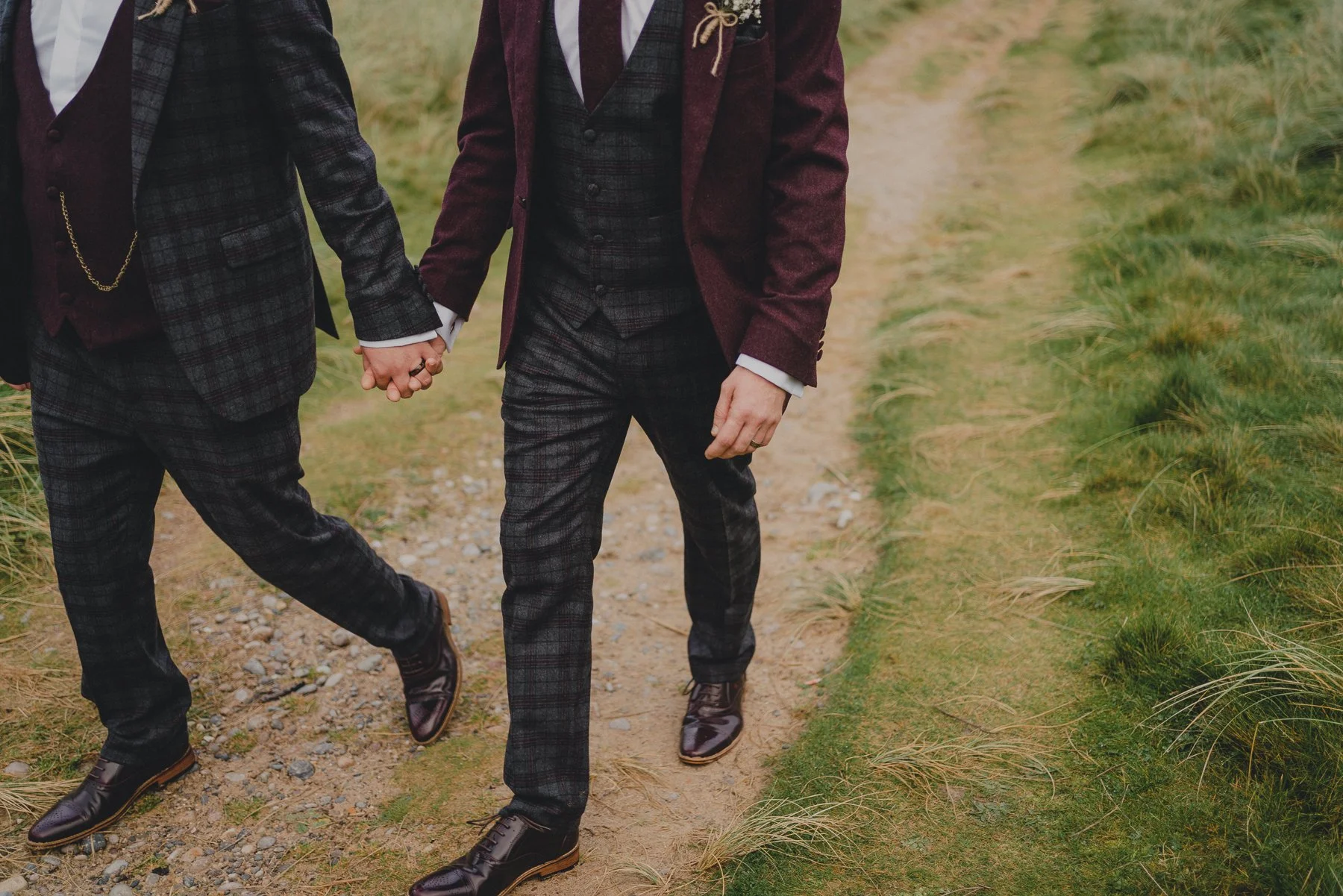 Close-up of two people holding hands while walking on a dirt path in a grassy outdoor area. They are dressed in formal plaid suits.