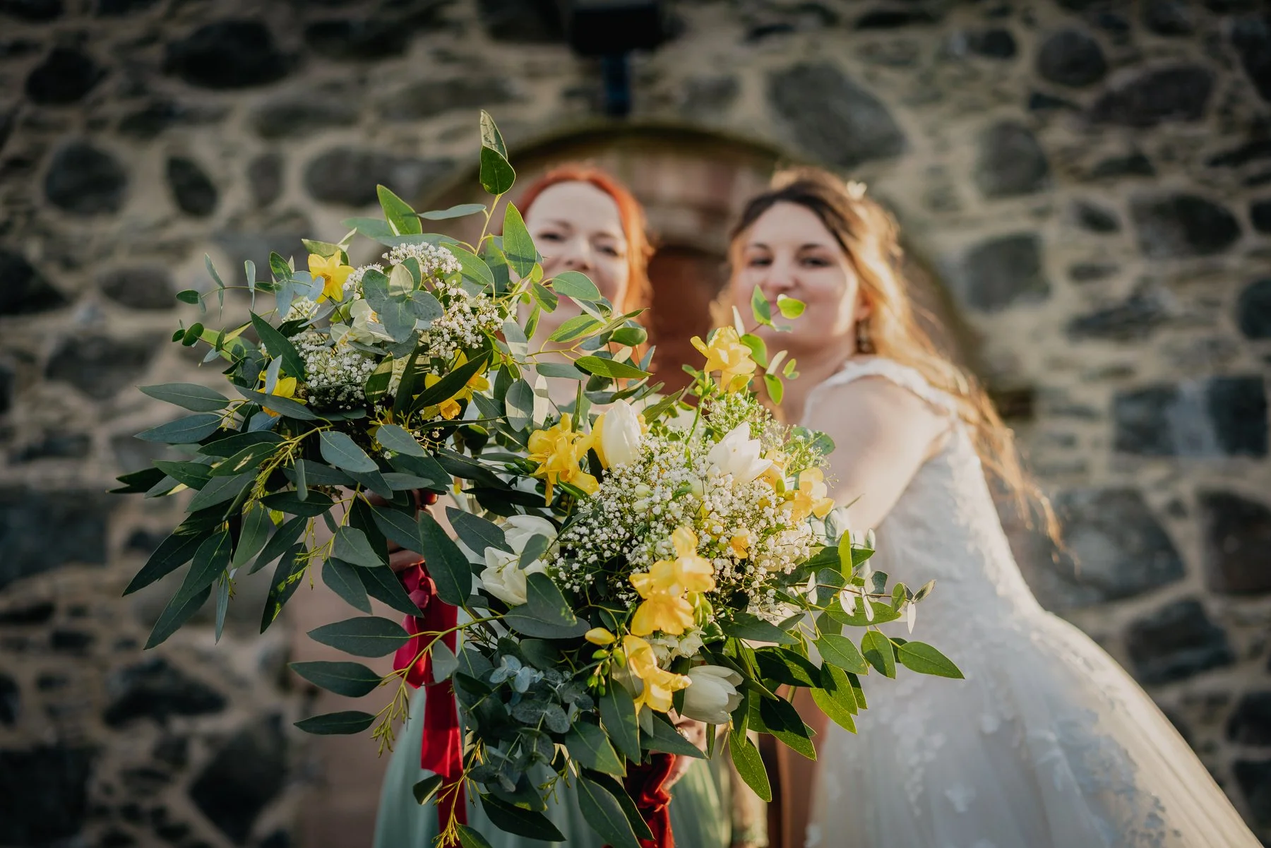 Bride holding a large bouquet of yellow and white flowers with greenery in front of a stone wall.