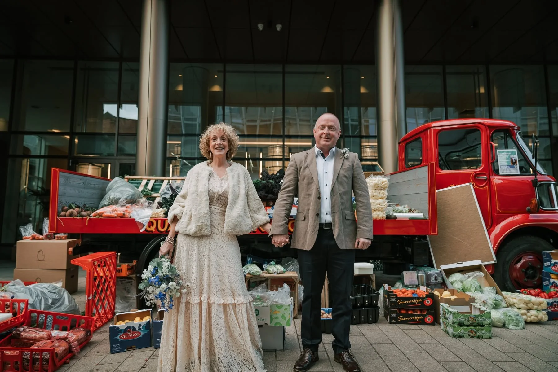 A happy couple in wedding attire standing and holding hands in front of a red truck selling fresh vegetables. The bride is wearing a vintage lace wedding dress with a faux fur stole, and the groom is in a beige suit with a boutonniere. They are outside a modern building with glass windows, surrounded by crates and boxes of produce like carrots, onions, and potatoes.