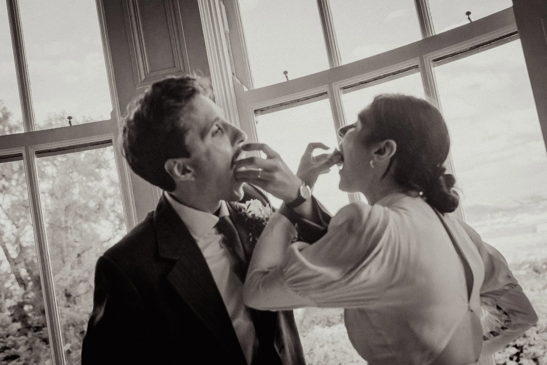 A man and a woman sharing a playful moment near large windows, with trees visible outside, in black and white.