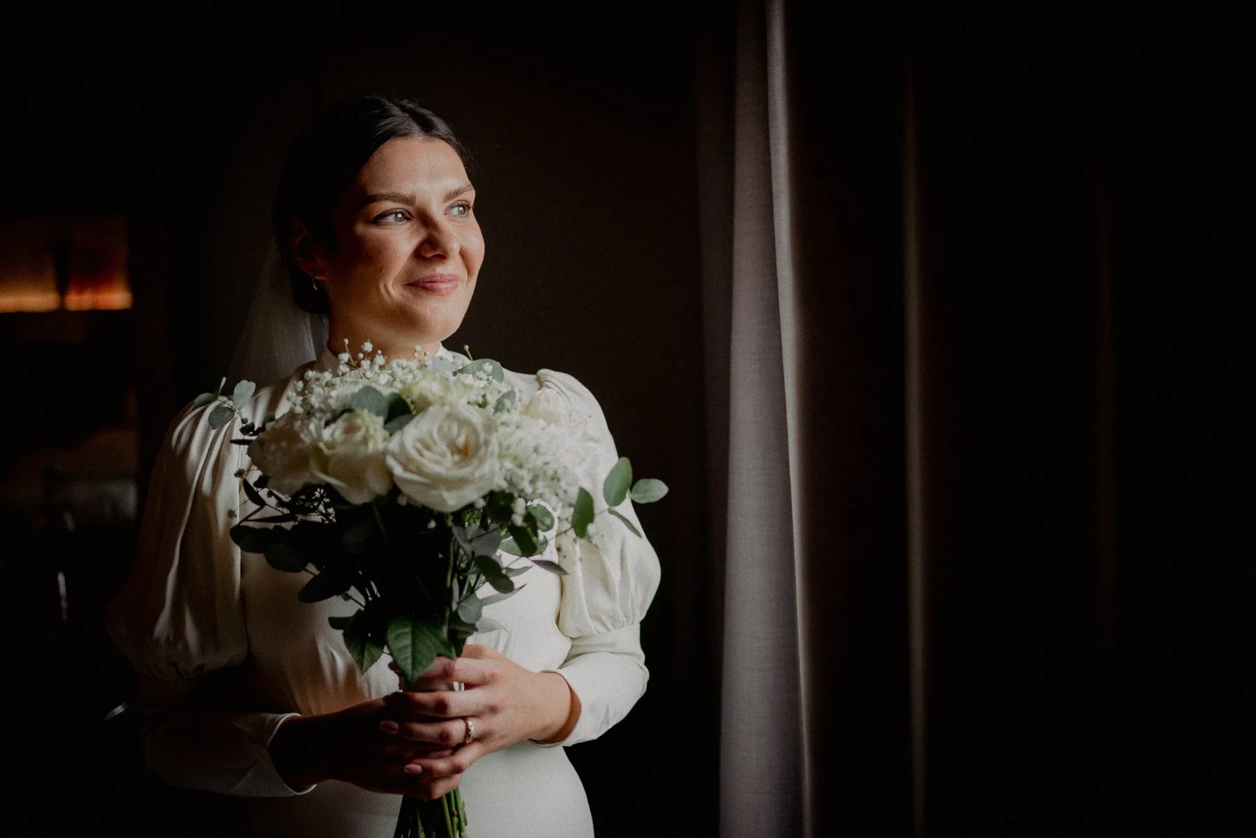 A woman in a white dress holding a bouquet of white flowers, standing near a window with dark curtains.