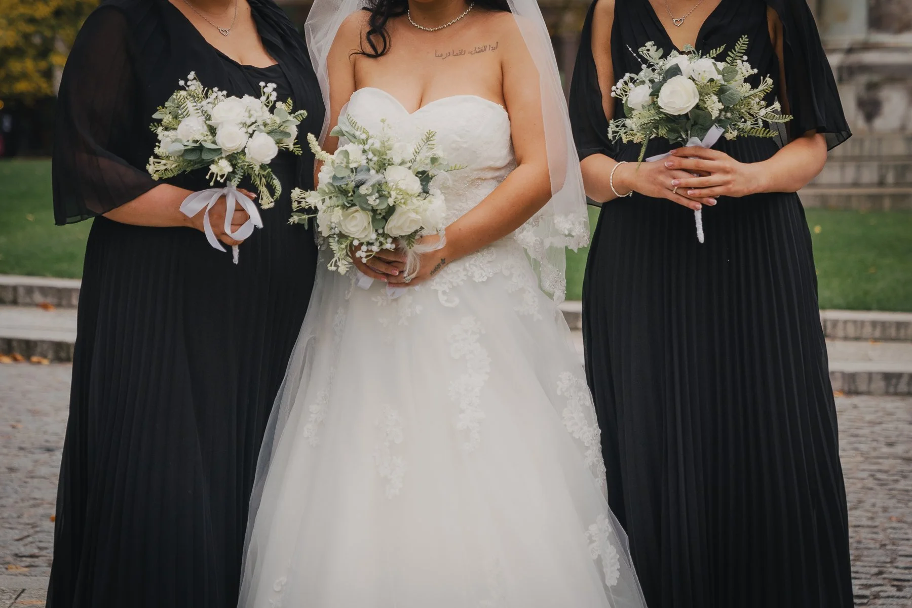 Close-up of a bride in a white wedding dress holding a bouquet of white roses, flanked by two women dressed in black with matching bouquets, outdoors with steps and grass in the background.