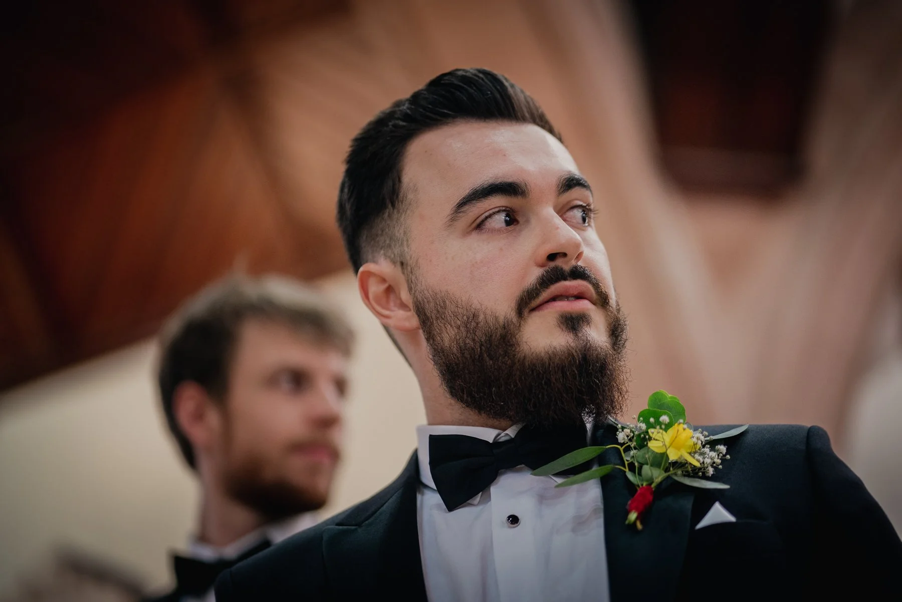 A man with dark hair and a beard wearing a tuxedo with a bowtie and a boutonniere, attending a formal event, with another man blurred in the background.