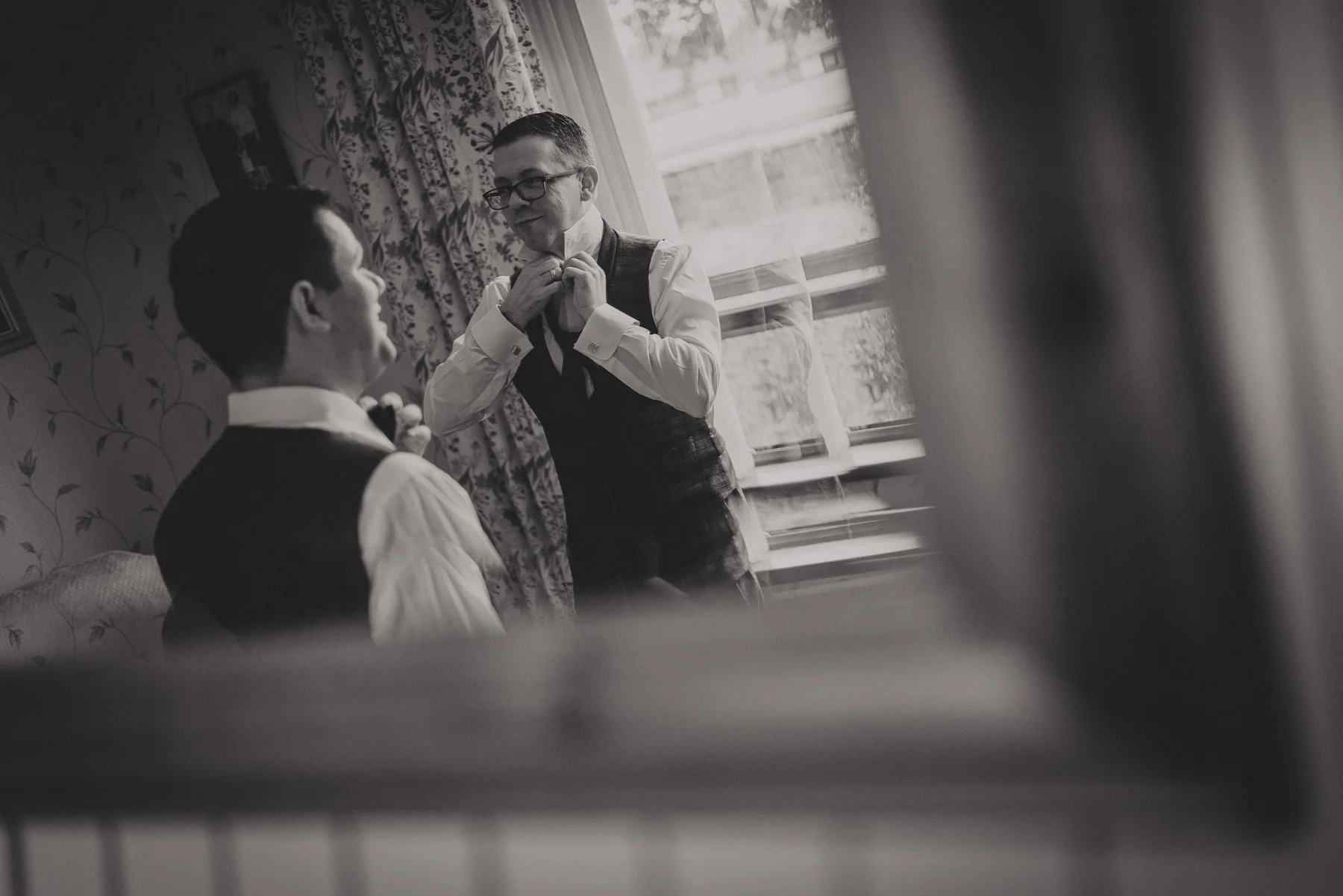 Black and white photo of two men in a room, one sitting and the other standing, adjusting a tie. The standing man is wearing glasses and a vest. The room has floral curtains and a window with a view outside.
