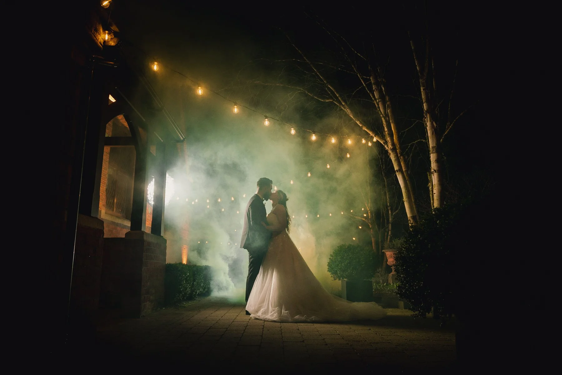 A couple in wedding attire sharing a kiss at night under string lights with trees and smoke in the background.