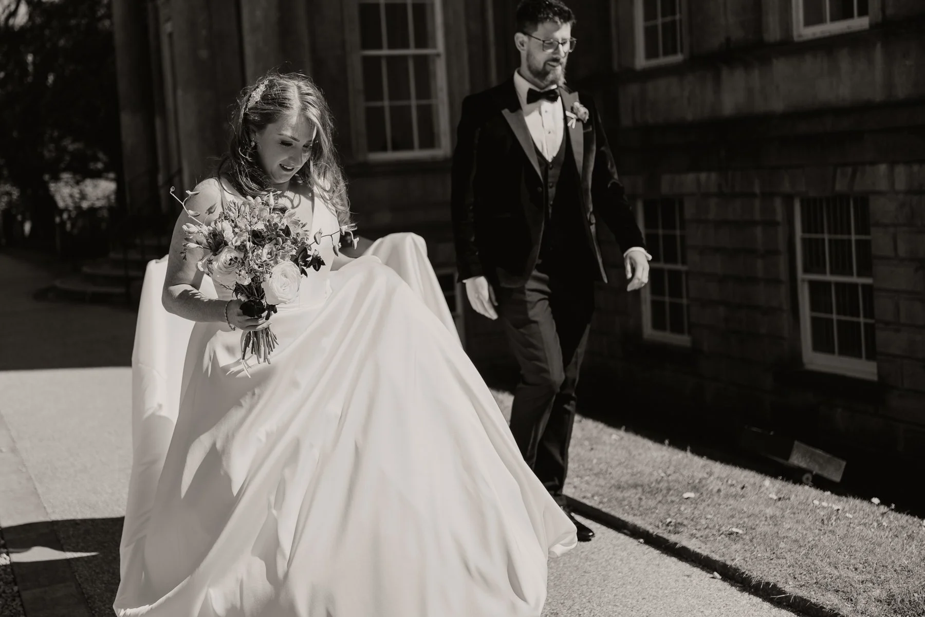 A bride holding a bouquet of flowers and lifting her wedding dress, walking beside a man in a tuxedo outside a building with brick walls.