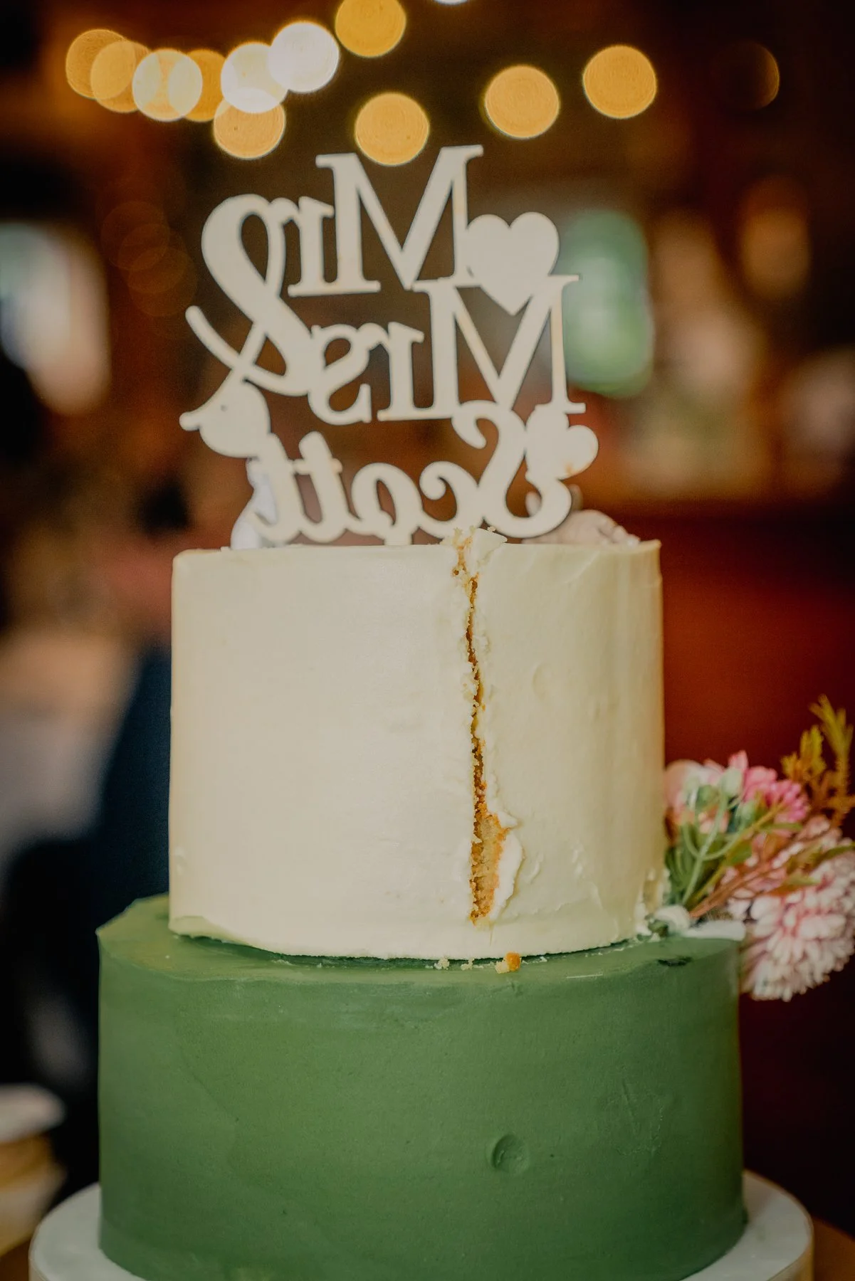 A wedding cake with a green bottom tier and a white top tier, decorated with a pink flower. A topper on the cake reads 'Mr & Mrs' with hearts and a cursive 'Love'.