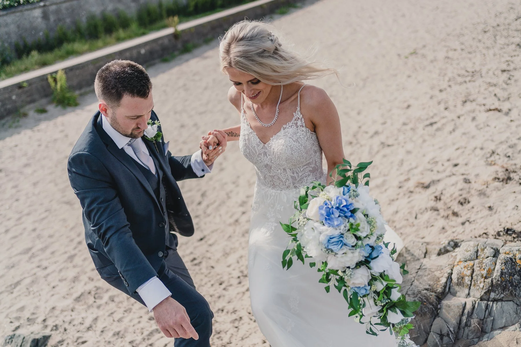 A bride and groom holding hands while walking on a sandy beach, with the bride carrying a bouquet of white and blue flowers and greenery.