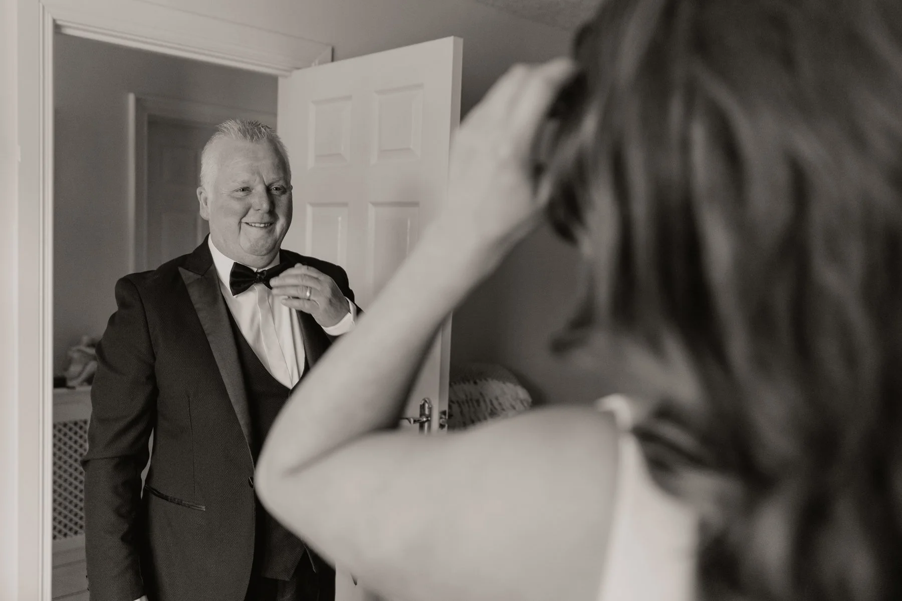 A man in a tuxedo adjusting his bow tie in front of a mirror, as a woman in the foreground touches her hair.
