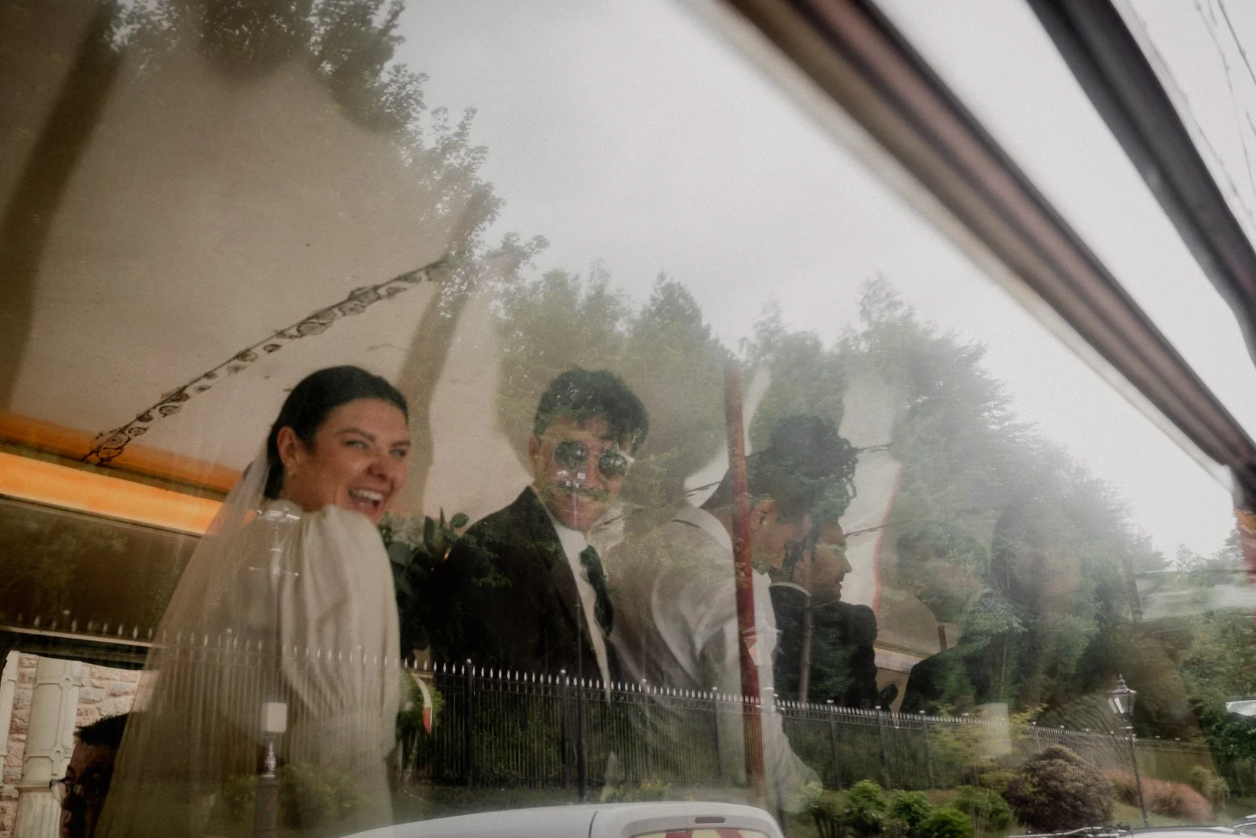 People looking through a car window, with the reflection showing trees and sky outside.