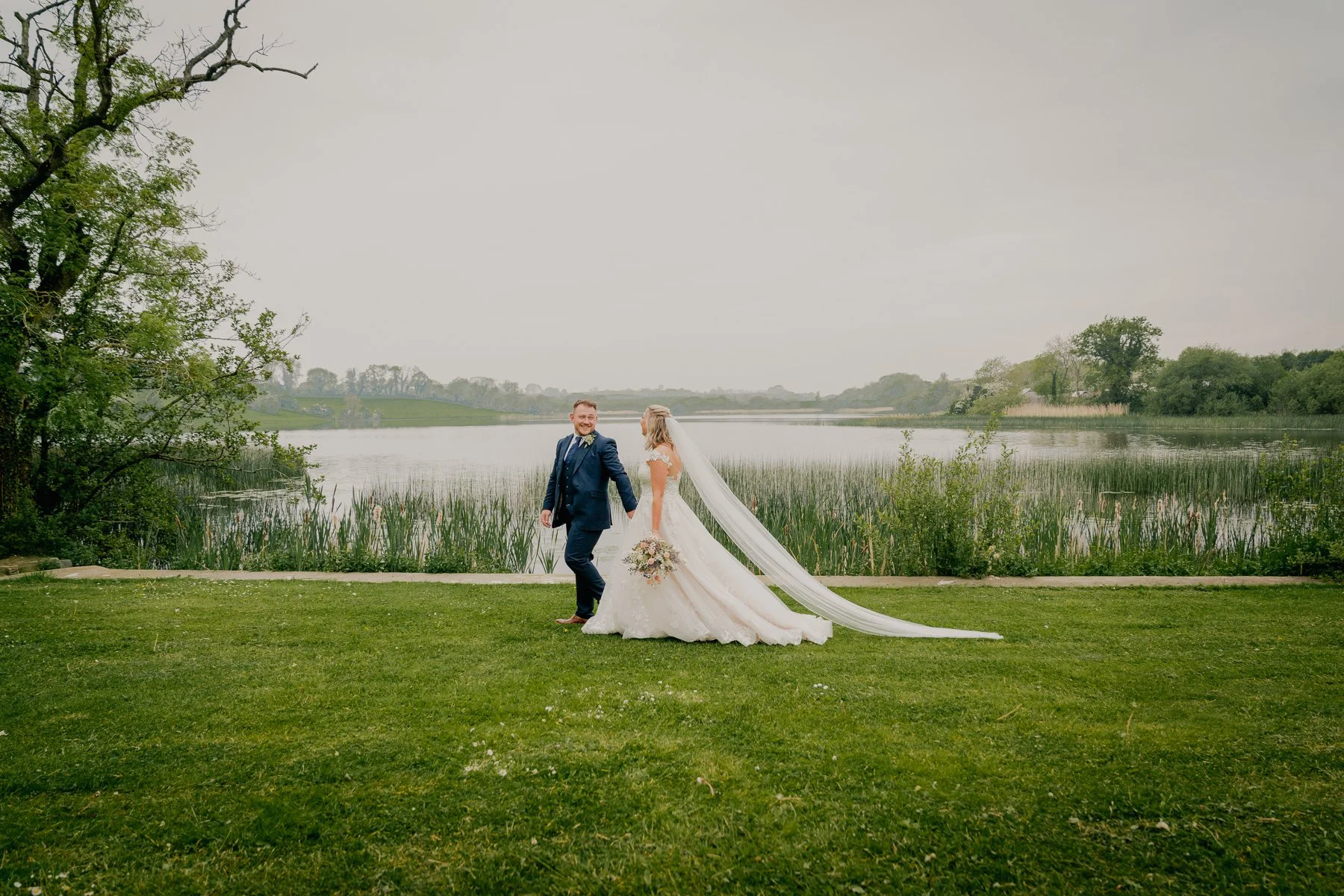 A bride and groom holding hands and smiling by a lake on their wedding day, with green grass, trees, and water in the background.
