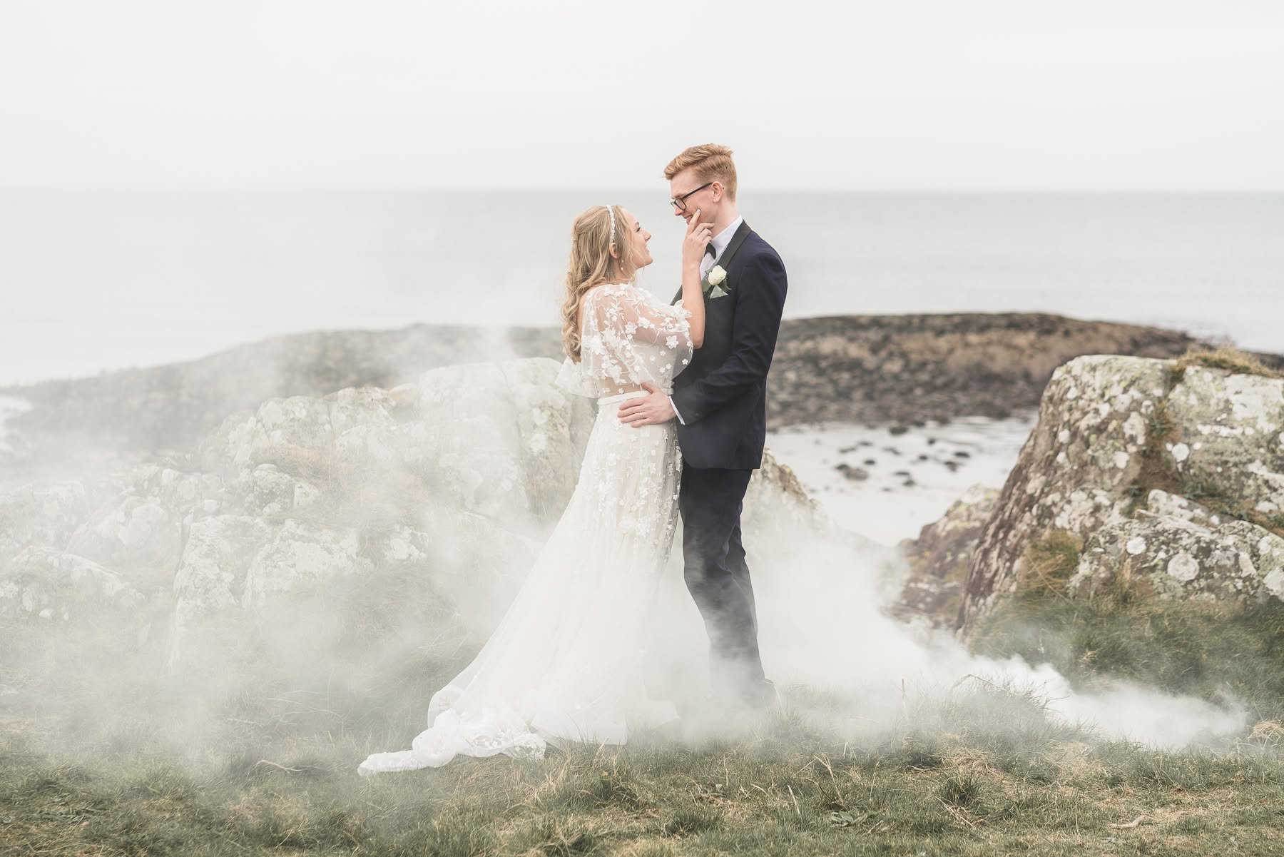 A bride and groom stand by rocky coast, smiling and holding each other in a romantic moment with foggy grass and sea in background.