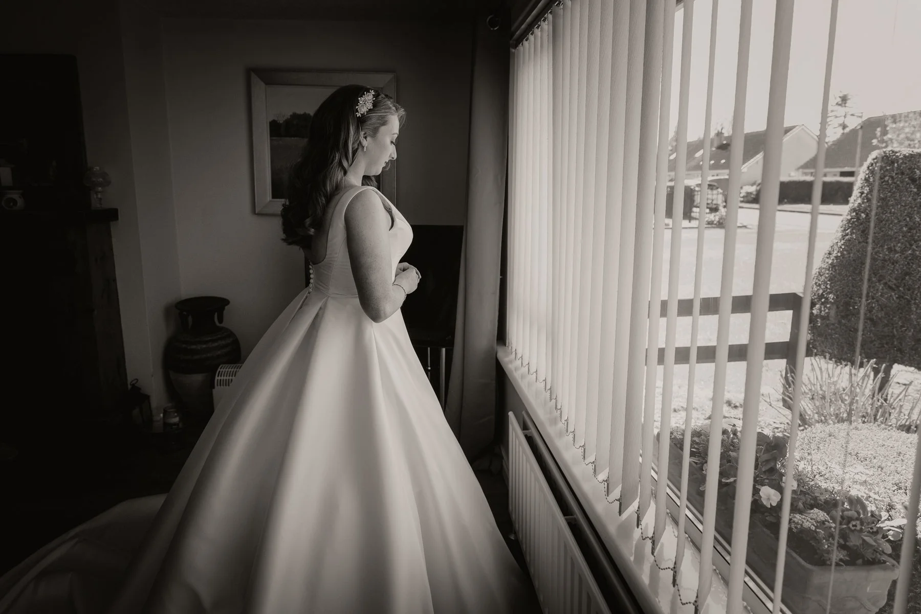 A bride in a white wedding gown with lace and buttons along the back, standing indoors by a window with vertical blinds, looking out at a suburban neighborhood with houses and a garden.