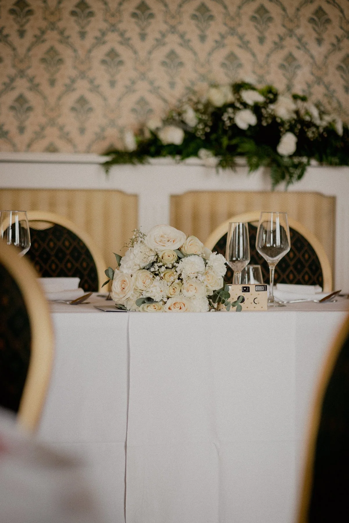 Elegant banquet table setting with a white floral centerpiece, wine glasses, and silverware in a decorated room.