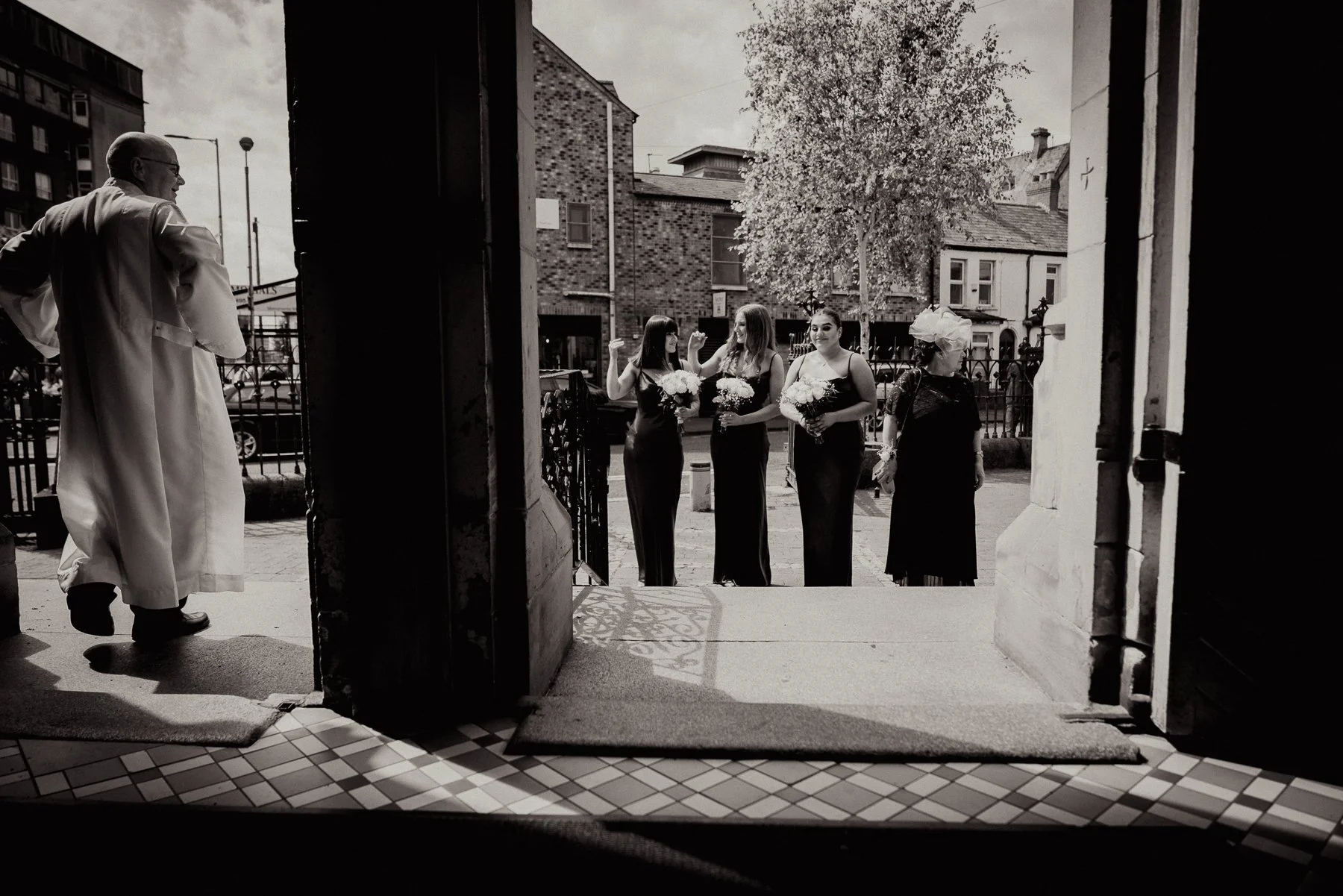 Black and white photo showing four women dressed in formal attire, holding bouquets, standing outside a building, engaged in conversation. An older man in a white coat is seen on the left side, standing with his hands on his hips, near an open door.