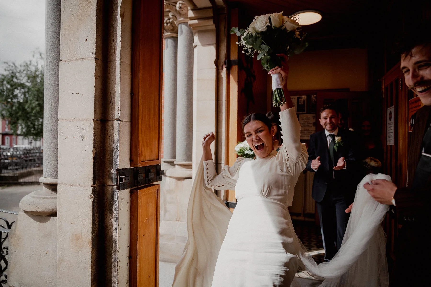 A bride celebrating joyfully with a bouquet of white roses inside a church, surrounded by smiling groomsmen, as she enters the venue.
