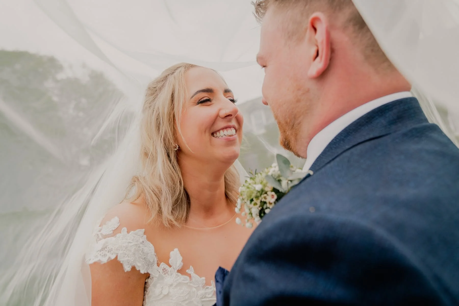 A newlywed couple standing close together under a transparent canopy, smiling happily, with the bride in a white wedding dress and the groom in a dark suit.