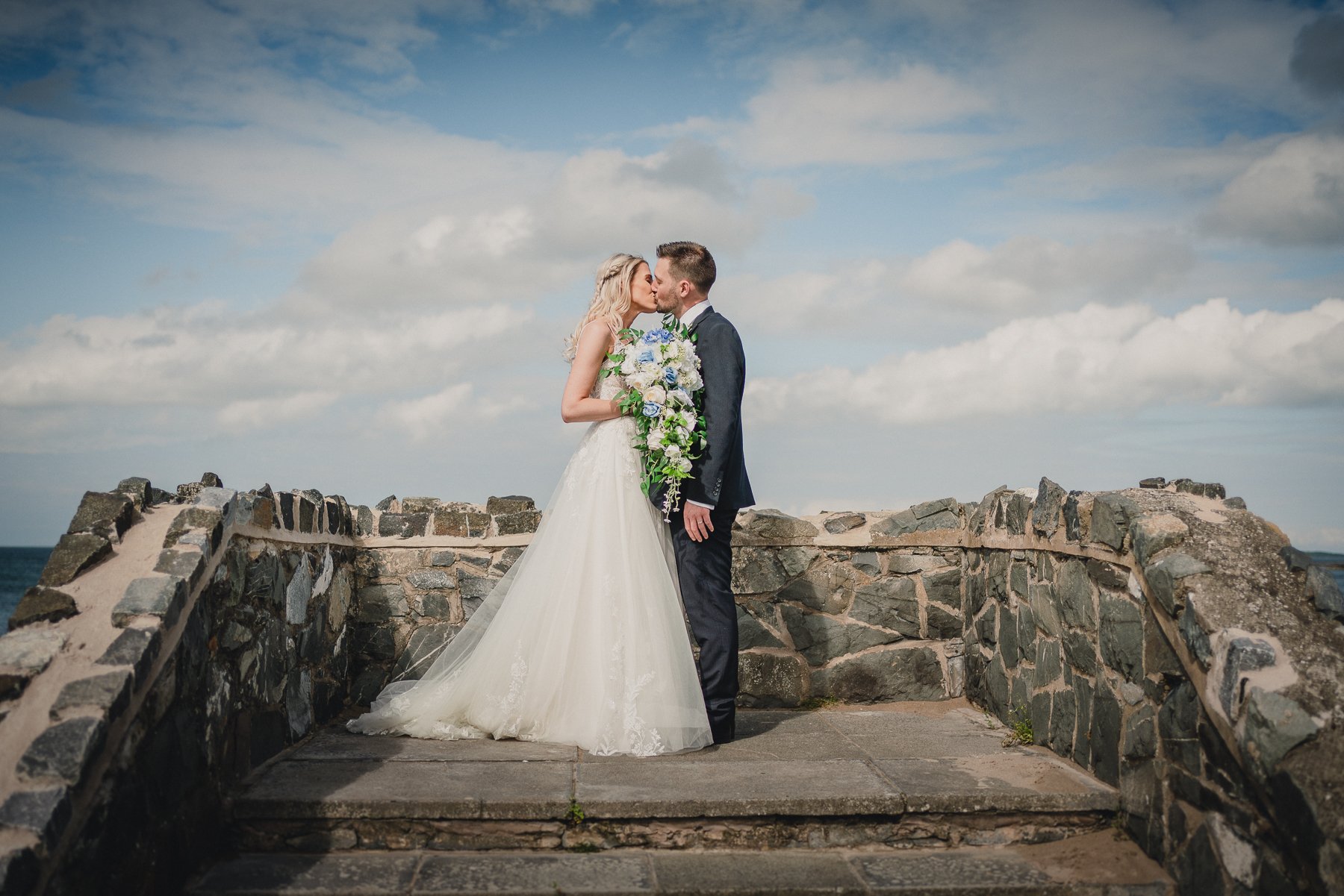 A bride and groom kissing on a stone bridge outdoors, with a cloudy sky and ocean in the background. The bride is holding a large bouquet of white and blue flowers.