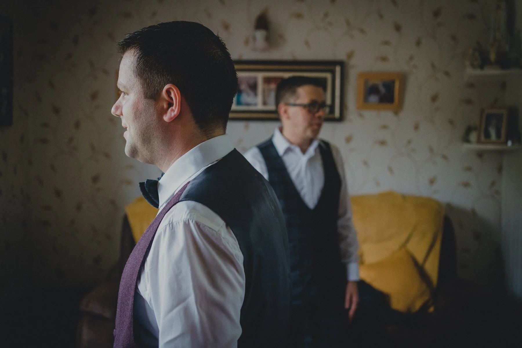 Two men dressed in formal attire, standing indoors with part of their profiles visible. The man in the foreground has short dark hair and wears a white shirt with a bow tie, vest, and sweater vest. The man in the background, wearing glasses, is sligh