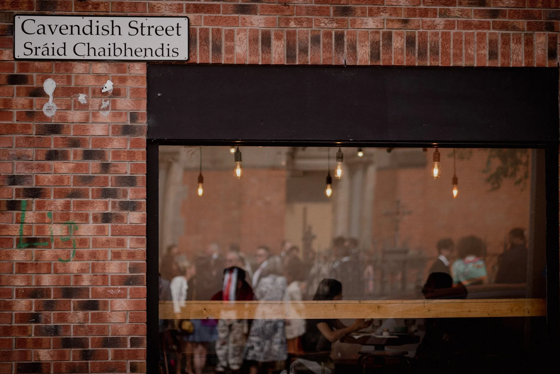 View through a window into a busy indoor space with hanging Edison-style light bulbs and several people mingling, with brick walls and a sign reading 'Cavendish Street' in English and Irish Gaelic.