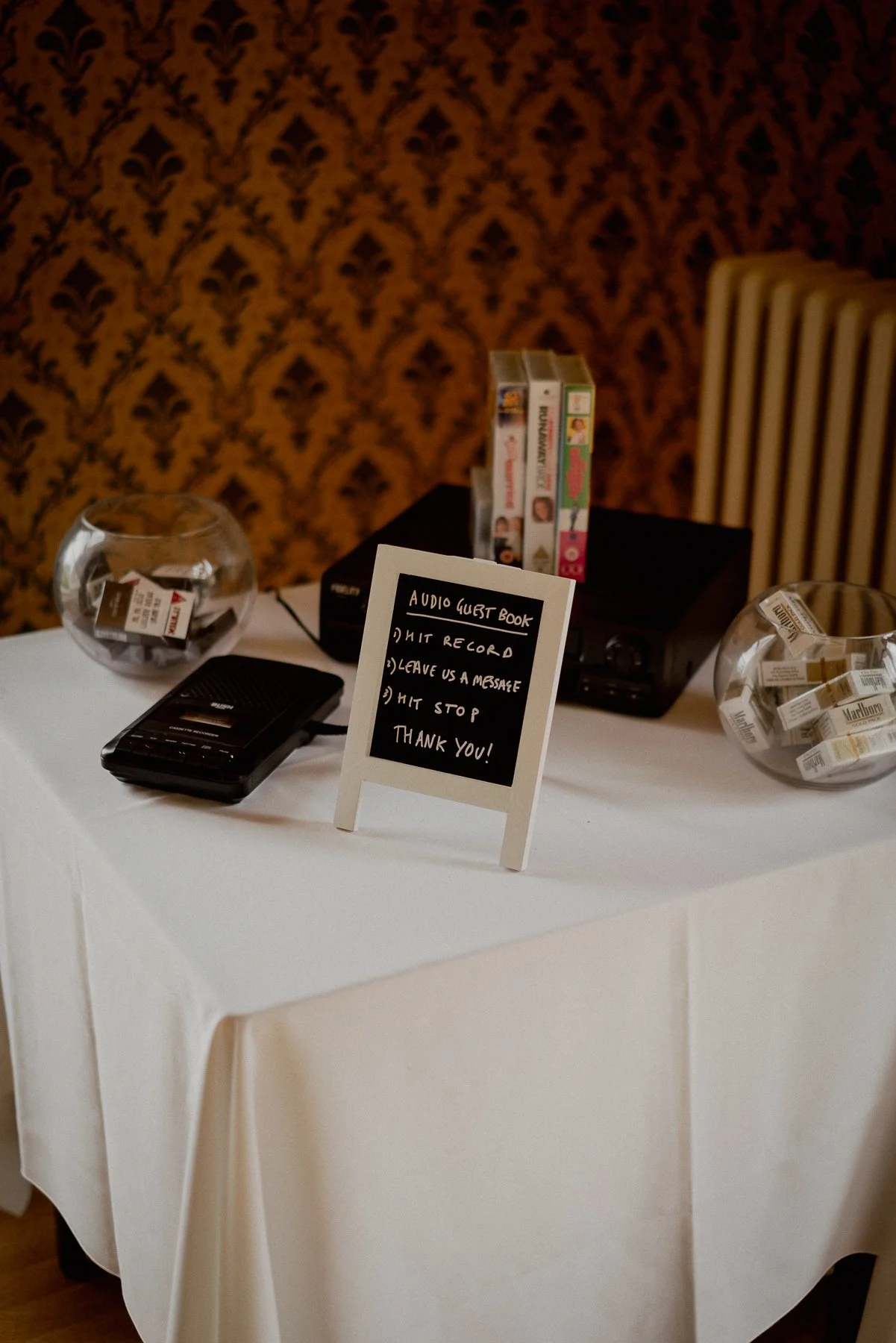 A table with two glass bowls filled with cigarette packs, a black audio recording device, and a small sign with instructions for an audio guest book on a white tablecloth. The background features patterned wallpaper and a chair with vertical stripes.