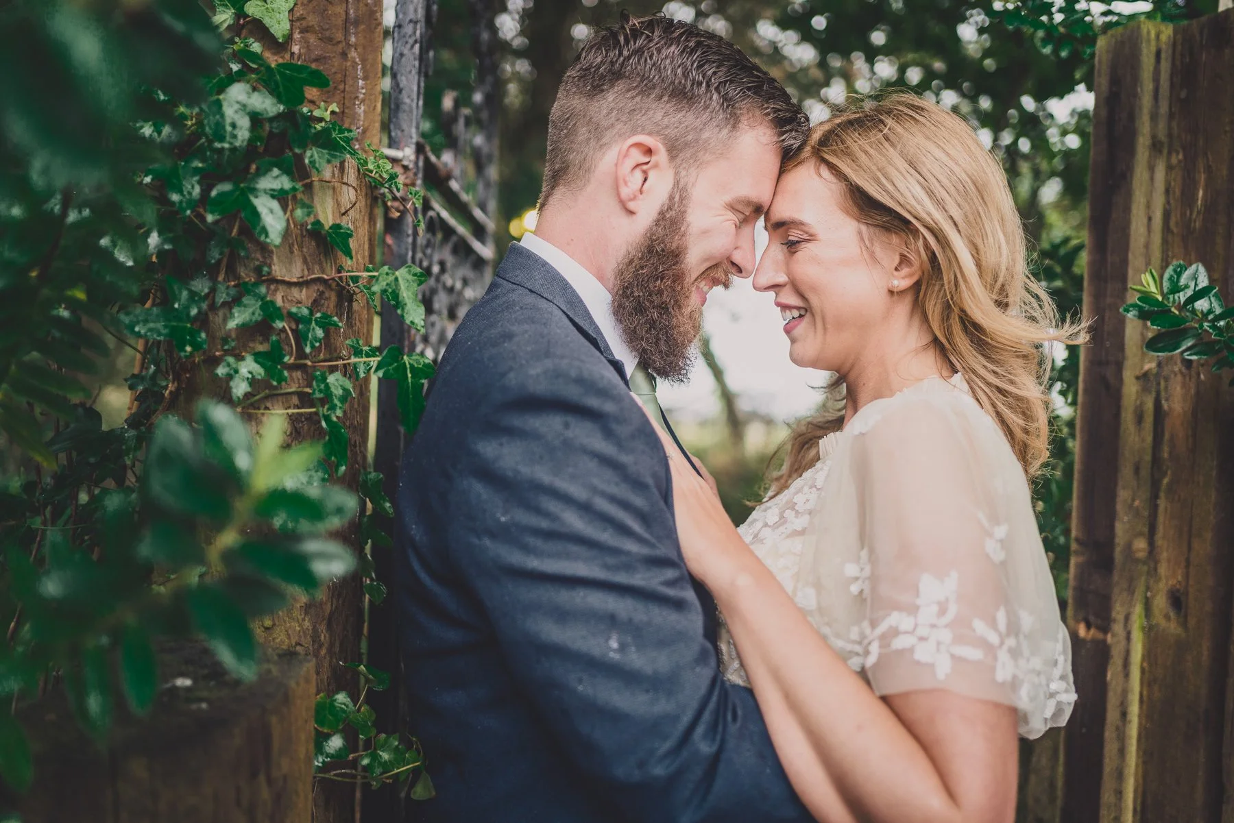 A smiling couple, a man with a beard and a woman with blonde hair, face to face outdoors among trees, touching foreheads, both dressed in formal attire, sharing a joyful moment.
