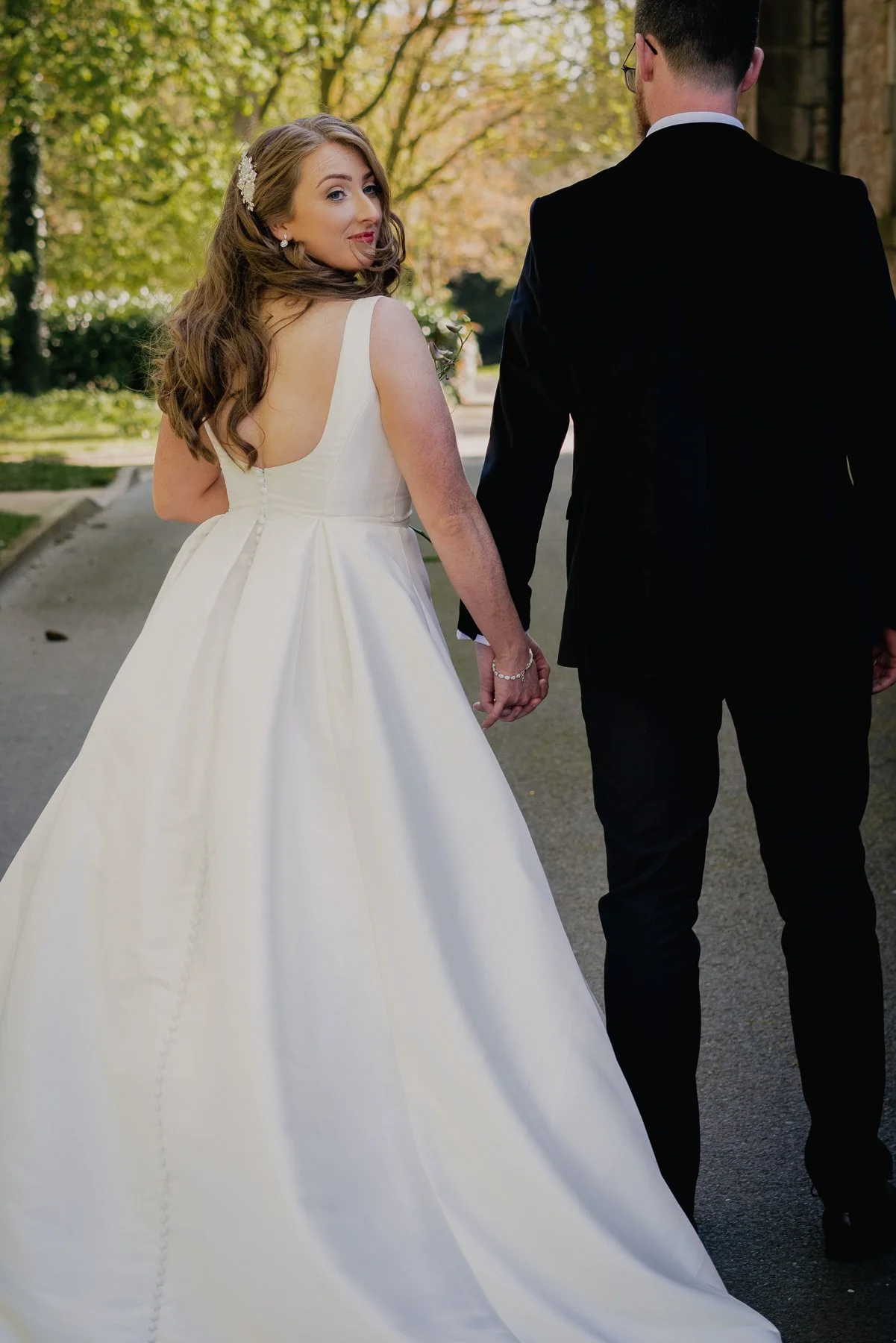 Bride and groom holding hands outdoors, bride in white wedding gown looking over her shoulder, groom in black suit with glasses, trees and a building in the background.