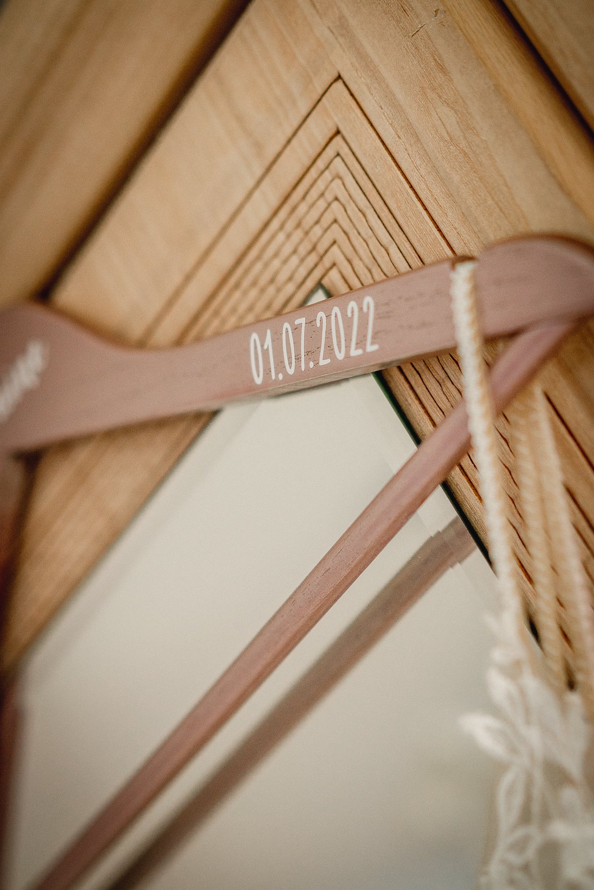 Close-up of a wooden structure with a pink ribbon that has the date 01.07.2022 written on it, and a part of a mirror and white lace seen in the background.