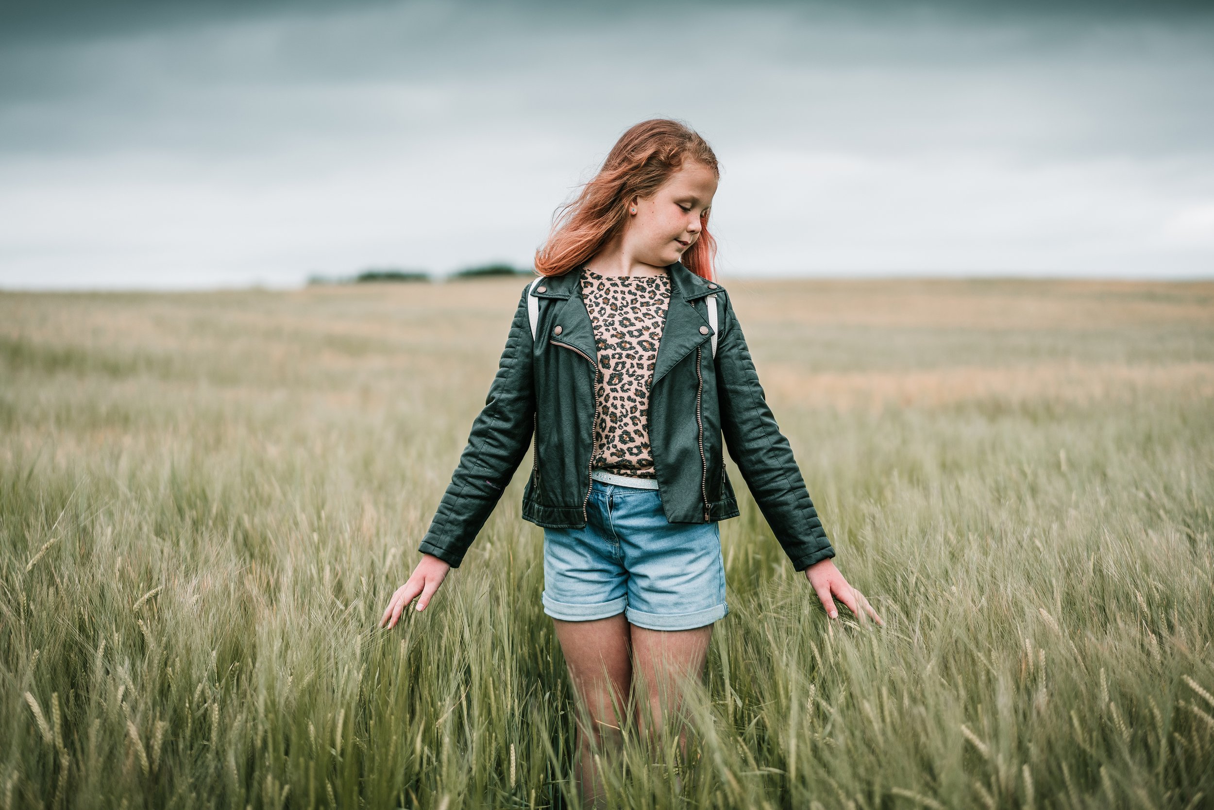 A young girl with red hair standing in a grassy field on a cloudy day, looking down and touching the grass with her hands.
