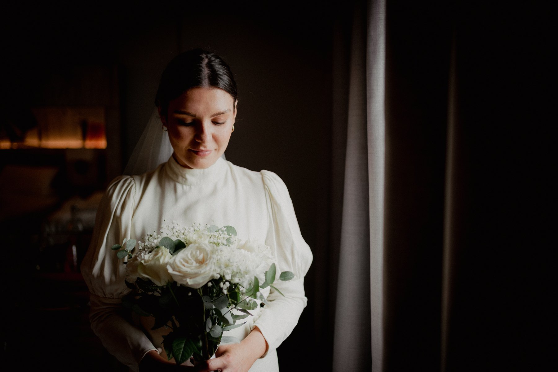 A woman in a white dress holding a bouquet of white flowers, standing near a window with curtains, with soft lighting.