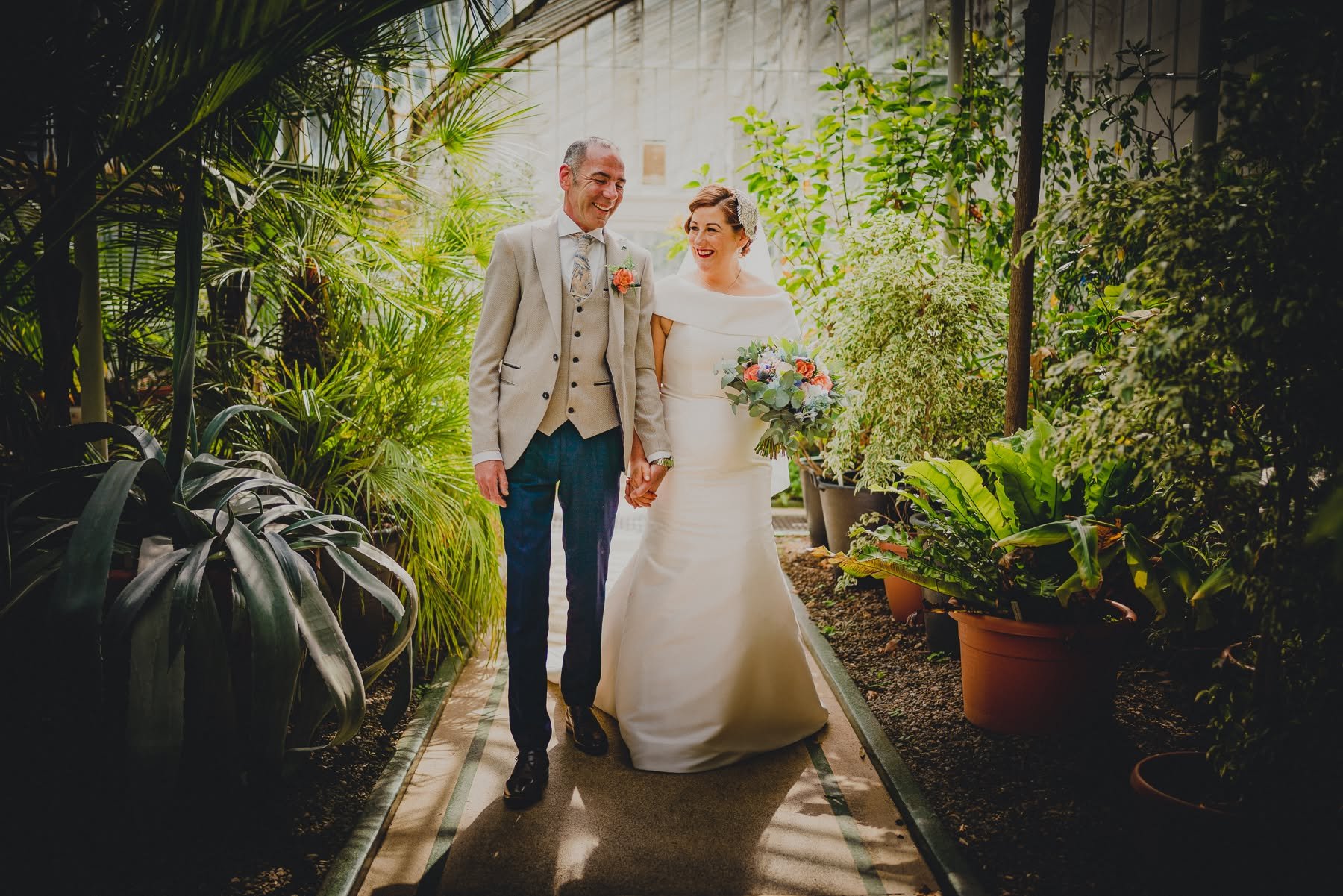 A bride and groom walking hand in hand through a lush greenhouse, surrounded by green plants and potted foliage, during their wedding celebration.