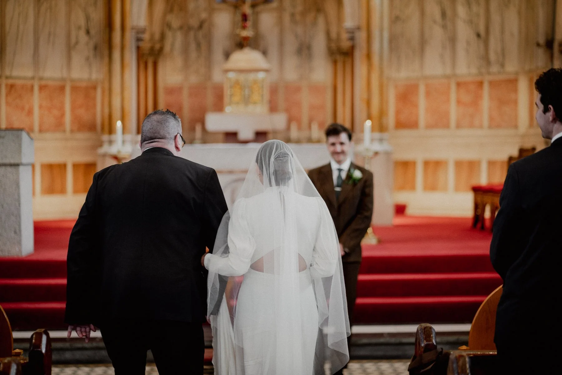 A bride and groom standing at the altar during a wedding ceremony inside a church, with a priest smiling in the background.