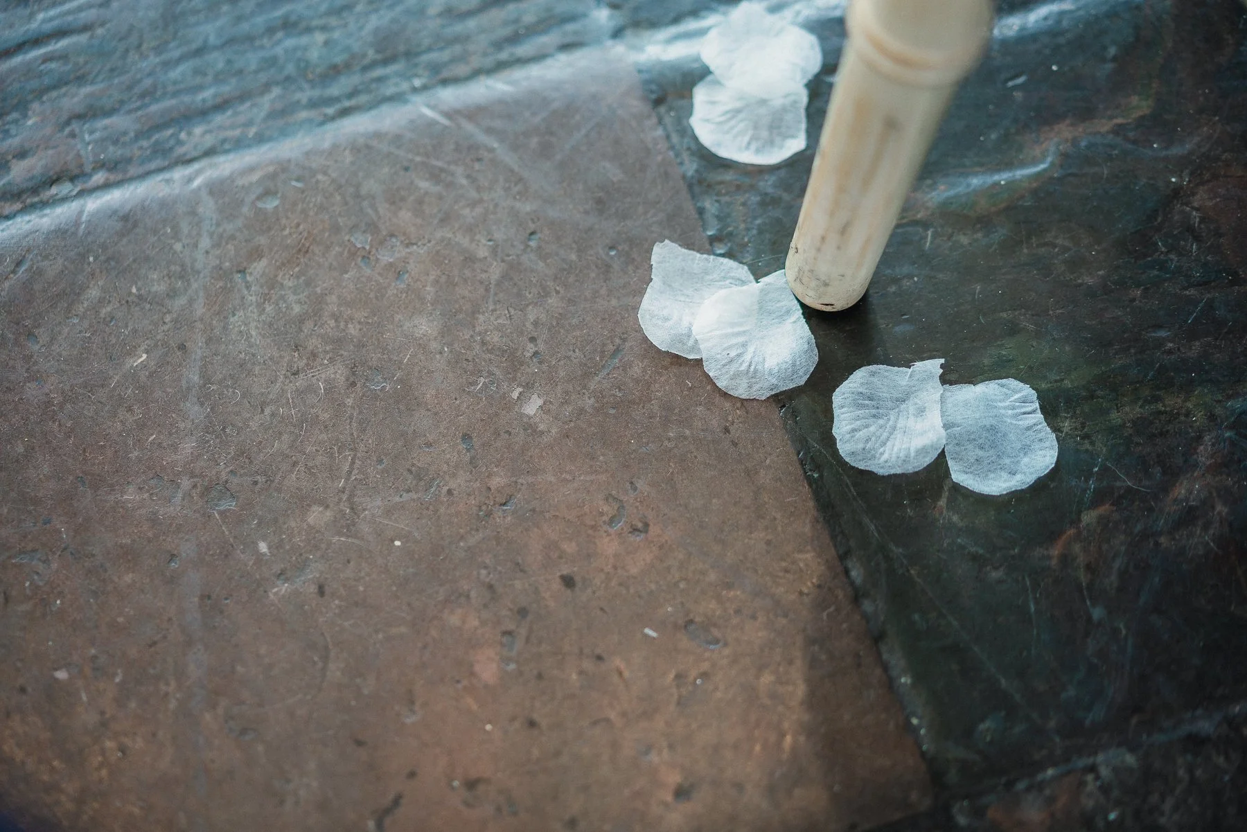 Close-up of white flower petals scattered on a tiled floor, with a light-colored cylindrical object leaning on a dark tile.
