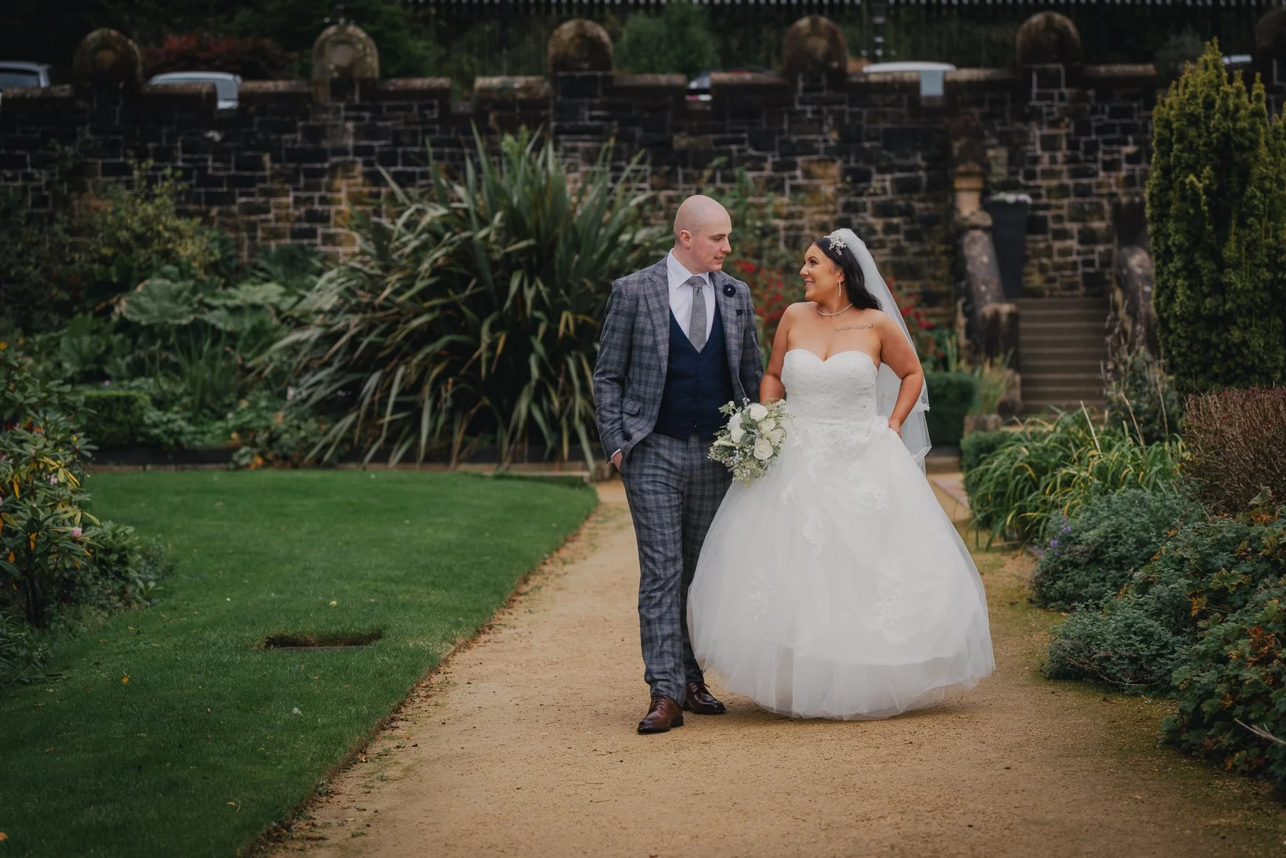 A newlywed couple walking together on a garden path. The bride is wearing a white strapless wedding gown and holding a bouquet of white flowers, and the groom is dressed in a gray plaid suit with a white shirt and tie. They are smiling at each other 