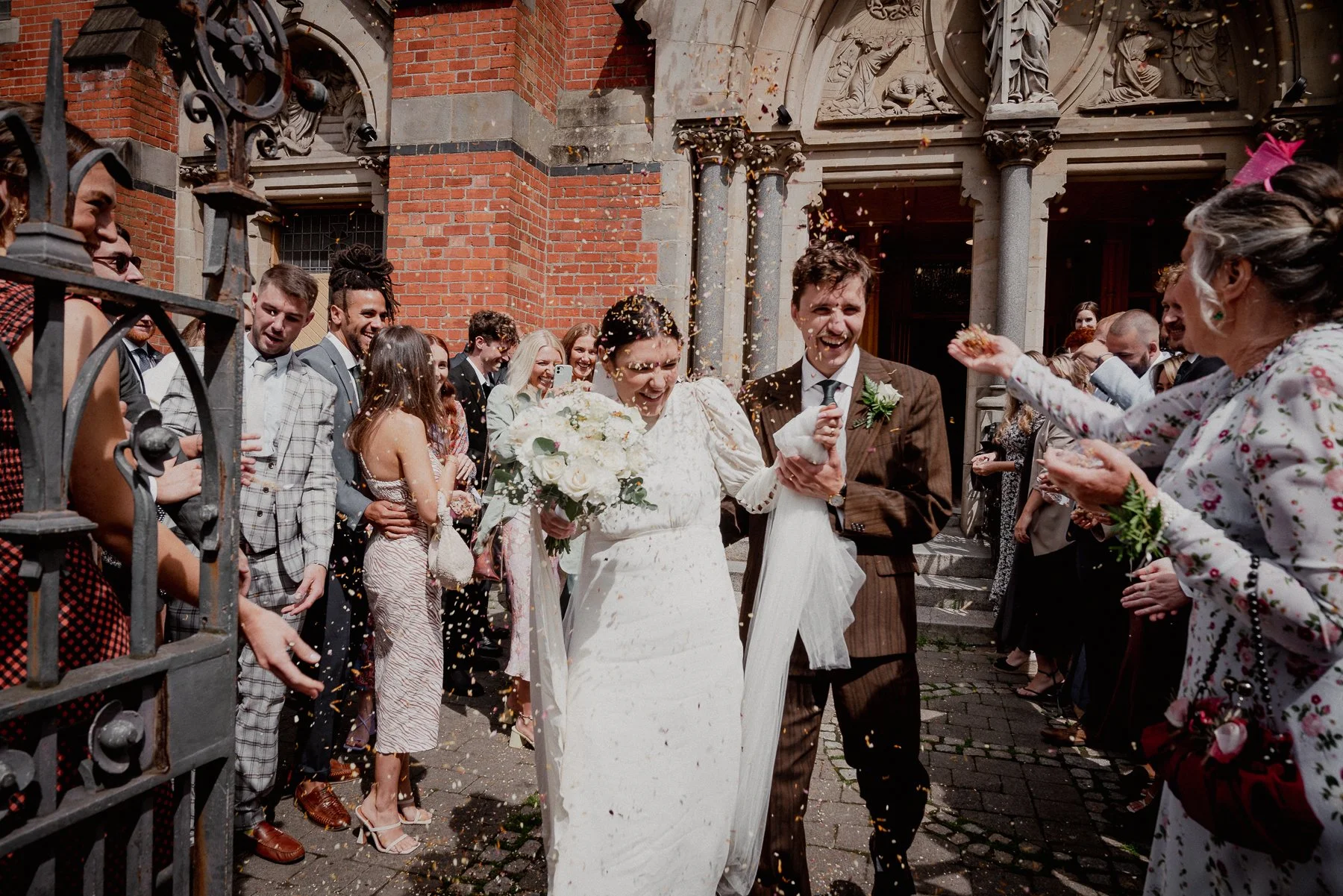 A wedding celebration with a bride and groom walking through a crowd outside a church, with guests throwing confetti and smiling.