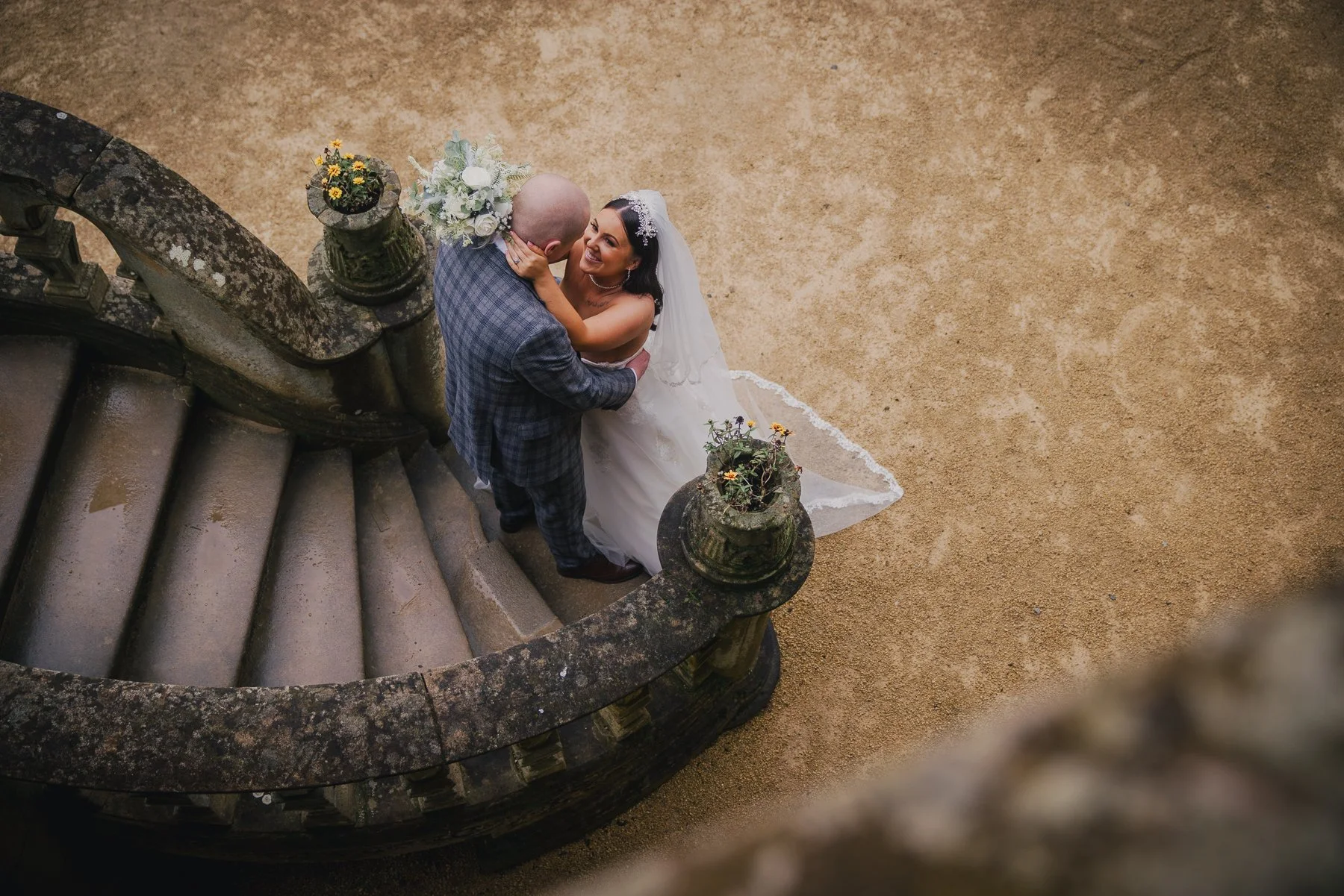 A bride and groom embrace on a curved stone staircase with potted plants at the top, viewed from above on a gravel surface.