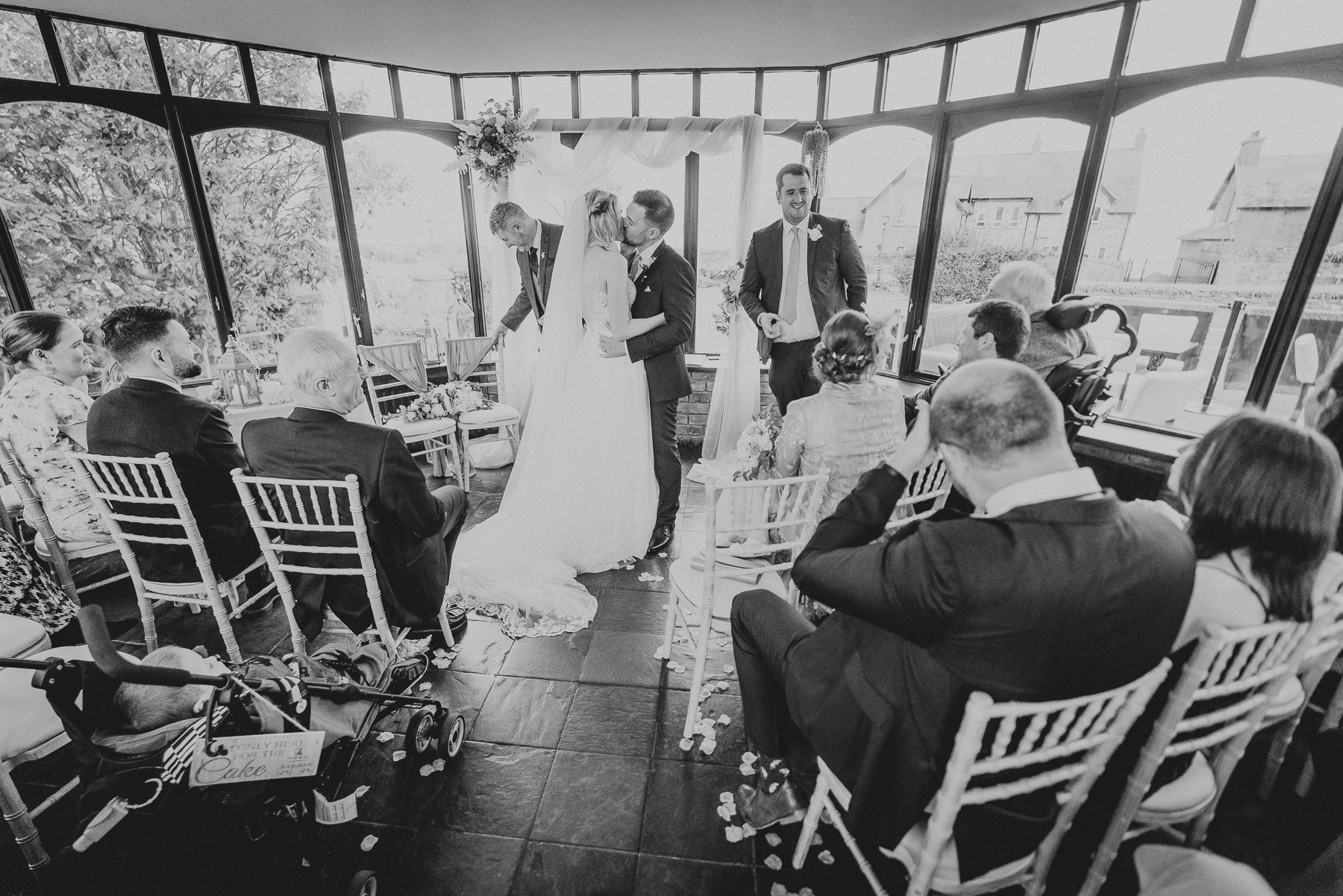 A black and white photograph of a wedding ceremony in a conservatory with large windows. The bride and groom are kissing, surrounded by seated guests, with floral decorations and draped fabric overhead. Guests are dressed formally, and a stroller wit