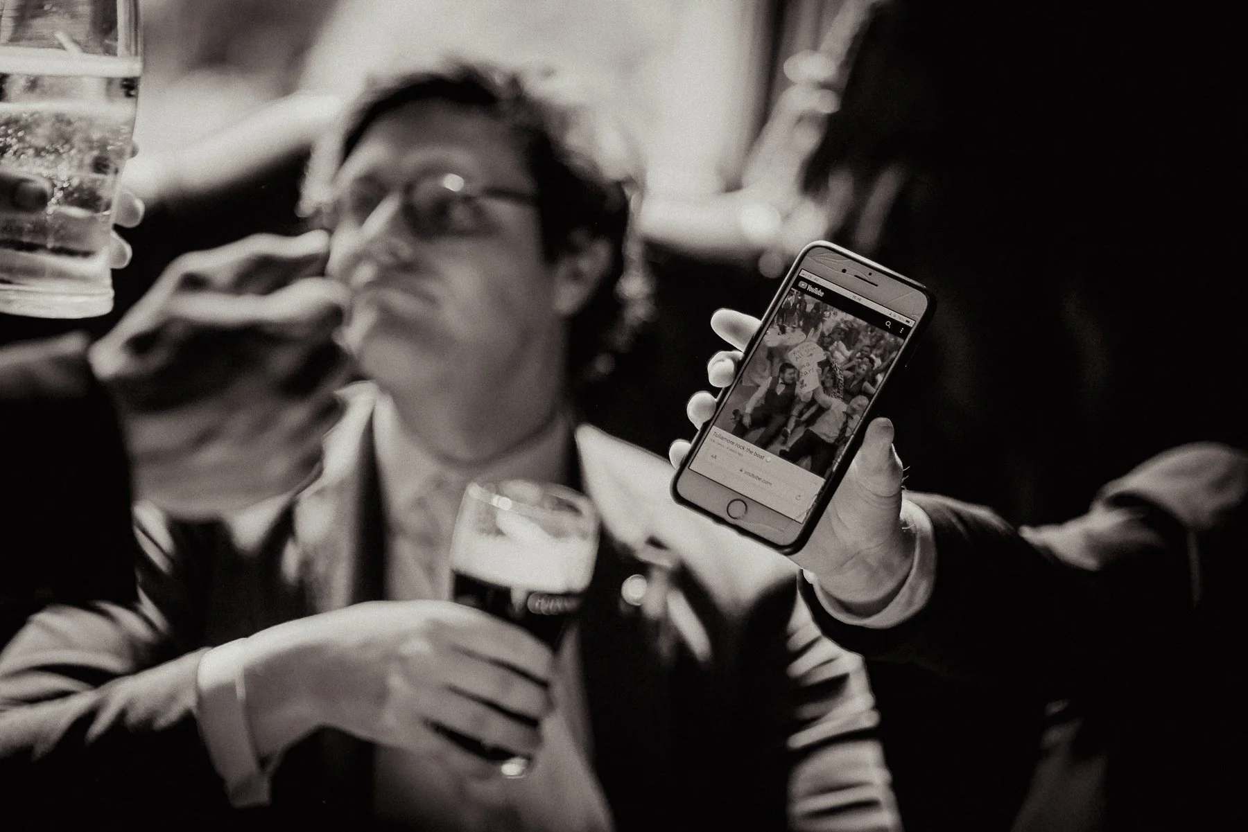 A person with glasses and long hair, seen in blurred focus, is holding a beer glass and a smartphone showing a photo of a crowd at a protest or gathering. The scene appears to be in a crowded indoor setting.