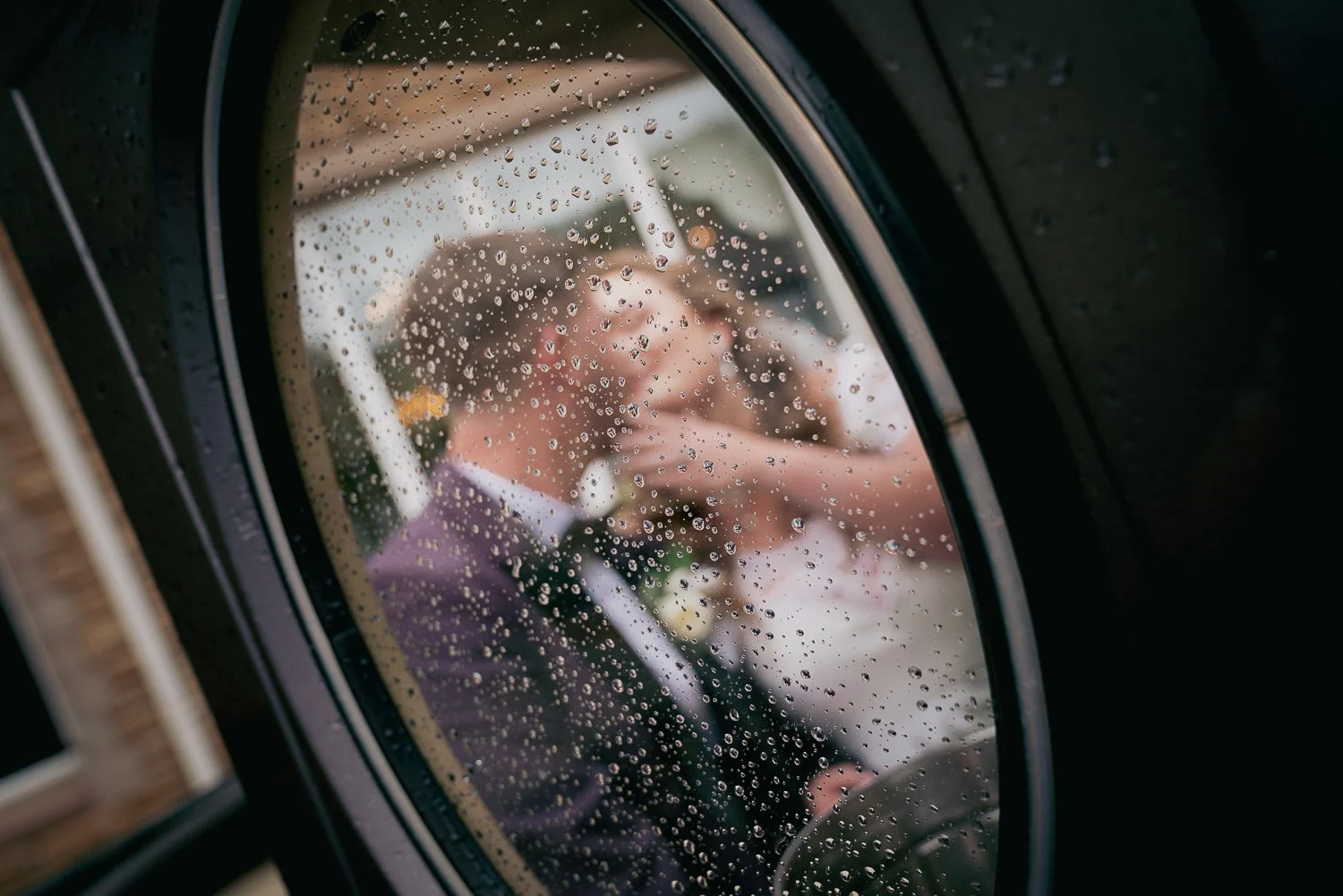 Reflected image of a bride and groom in a rain-covered window, with the rain drops clearly visible on the glass.