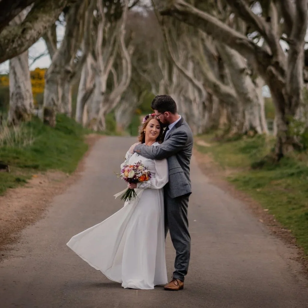 A newlywed couple hugging on a dirt road surrounded by a tunnel of twisted, leafless tree branches, with the bride holding a colorful bouquet and in a white dress, and the groom in a gray suit.