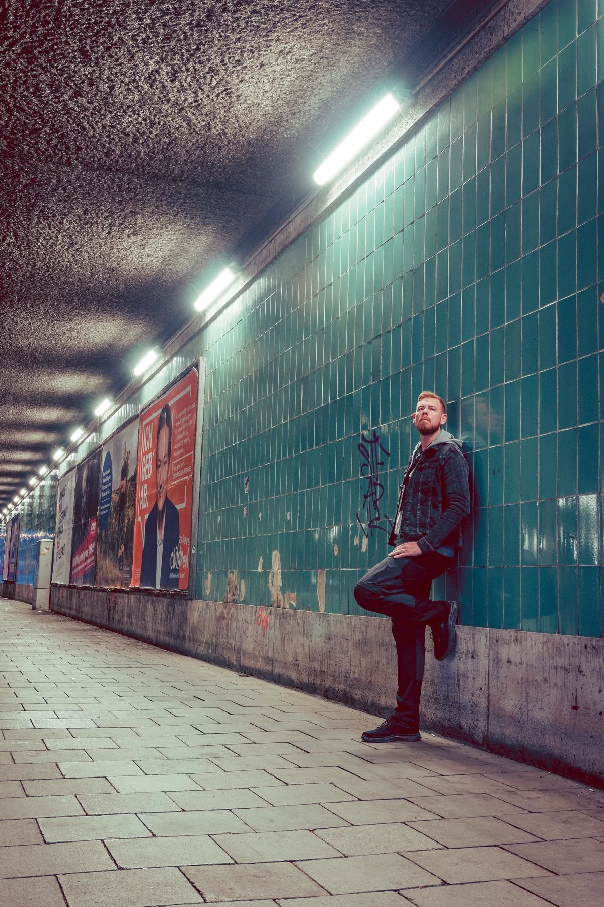 A man with a beard, wearing a denim jacket and black pants, leaning against a green tiled wall in an underground tunnel with advertisements, fluorescent lighting, and a tiled walkway.