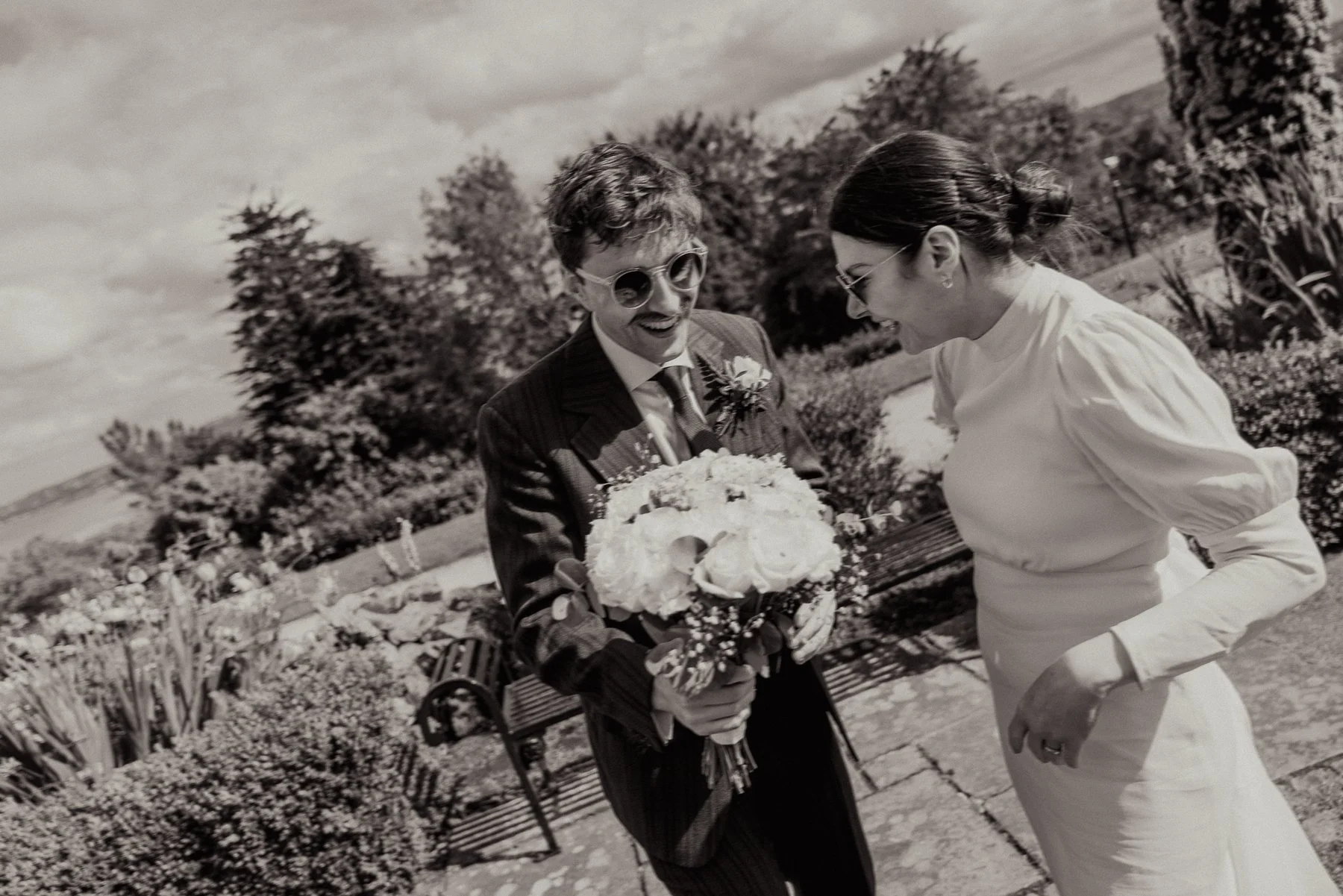 A man in a suit holding a bouquet of flowers, smiling, with a woman in a white dress smiling back, outdoors in a garden with plants and trees.