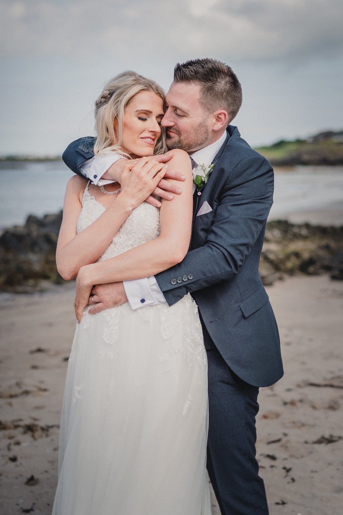 A bride and groom hugging on a beach, with water and rocks in the background, the bride wearing a white lace wedding dress and the groom in a dark suit.