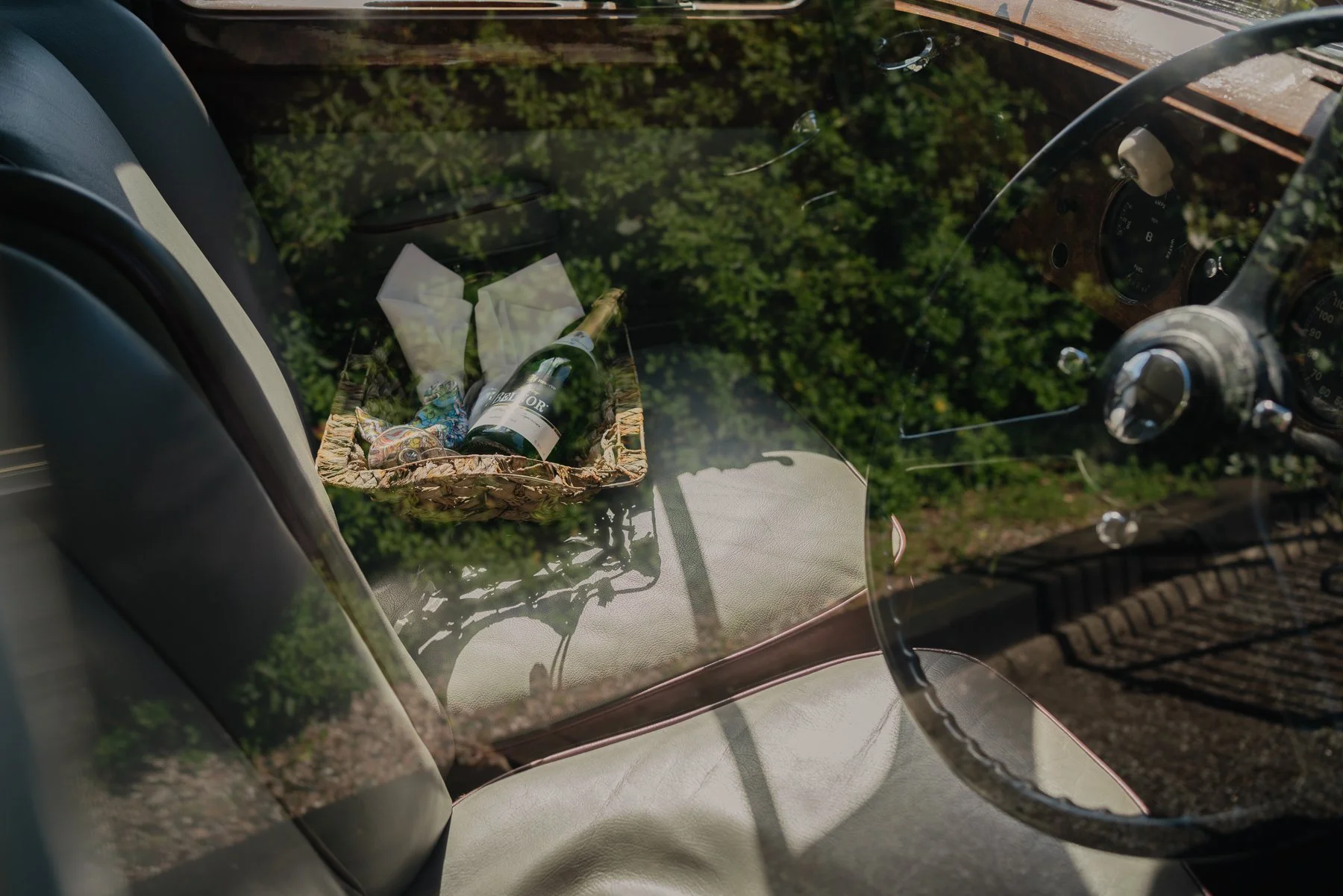 The image shows a view through the front windshield of a vehicle, reflecting trees and sky outside. Inside, a small wicker basket on the dashboard contains a bottle of wine, two wine glasses, some wrapped items, and a fabric item. The vehicle's inter