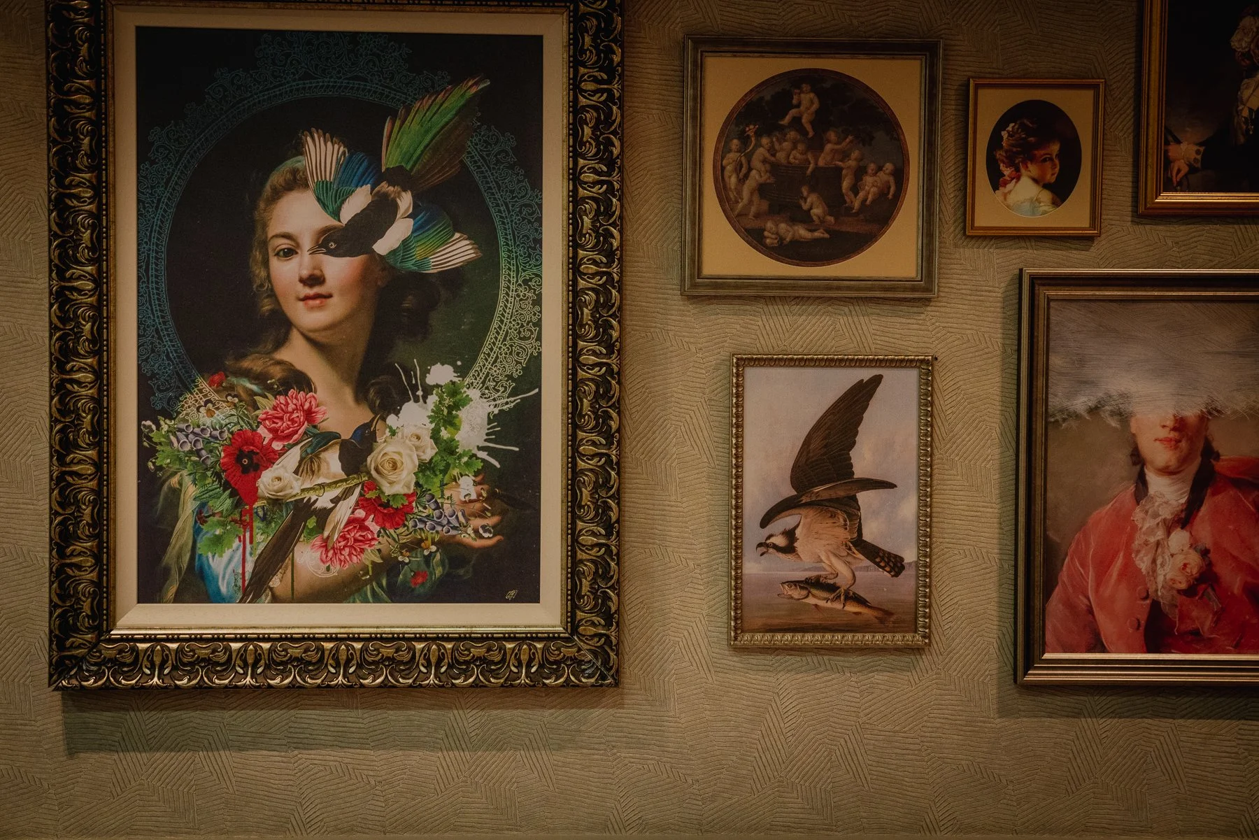 Wall of framed artwork including a prominent surreal portrait of a woman with colorful birds and flowers, surrounded by smaller paintings of classical, portrait, and bird images.