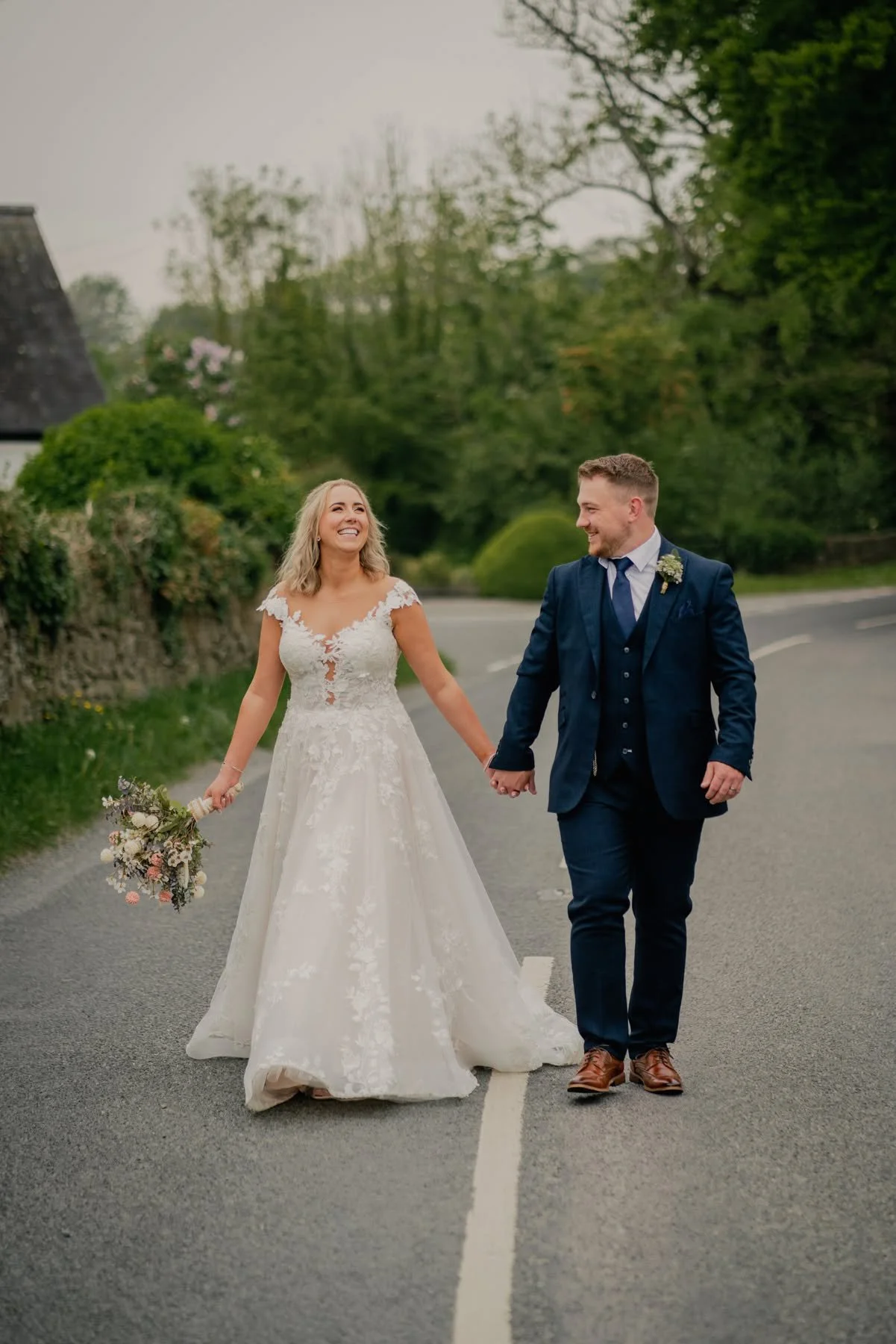 A smiling bride and groom walking hand in hand across an empty road on their wedding day, with the bride holding a bouquet of flowers and dressed in a white lace wedding gown, and the groom in a dark blue suit with a boutonniere, outdoors with trees and a cloudy sky in the background.
