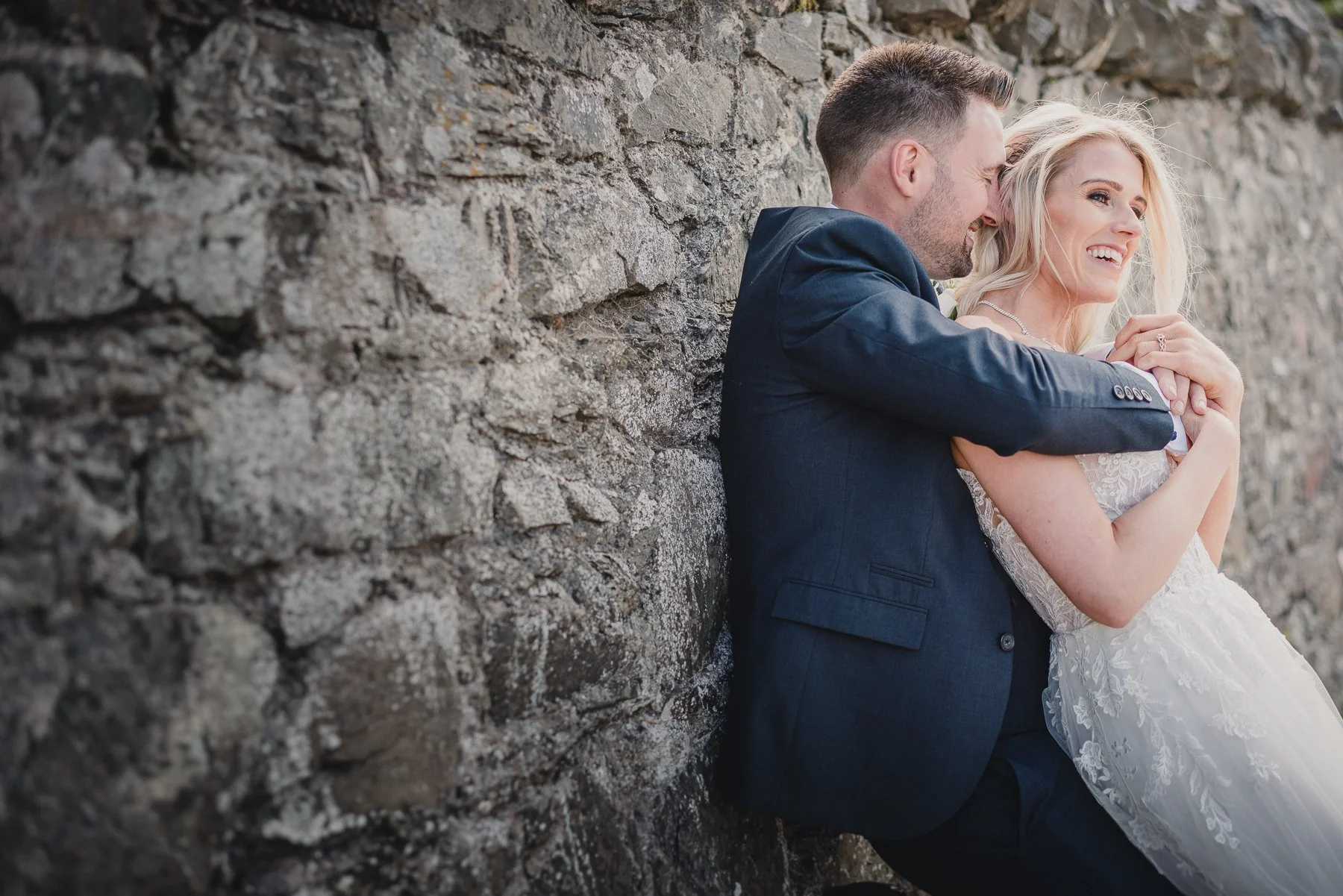 A smiling newlywed couple leaning against a stone wall, with the groom in a dark suit and the bride in a white wedding dress, embracing each other affectionately.