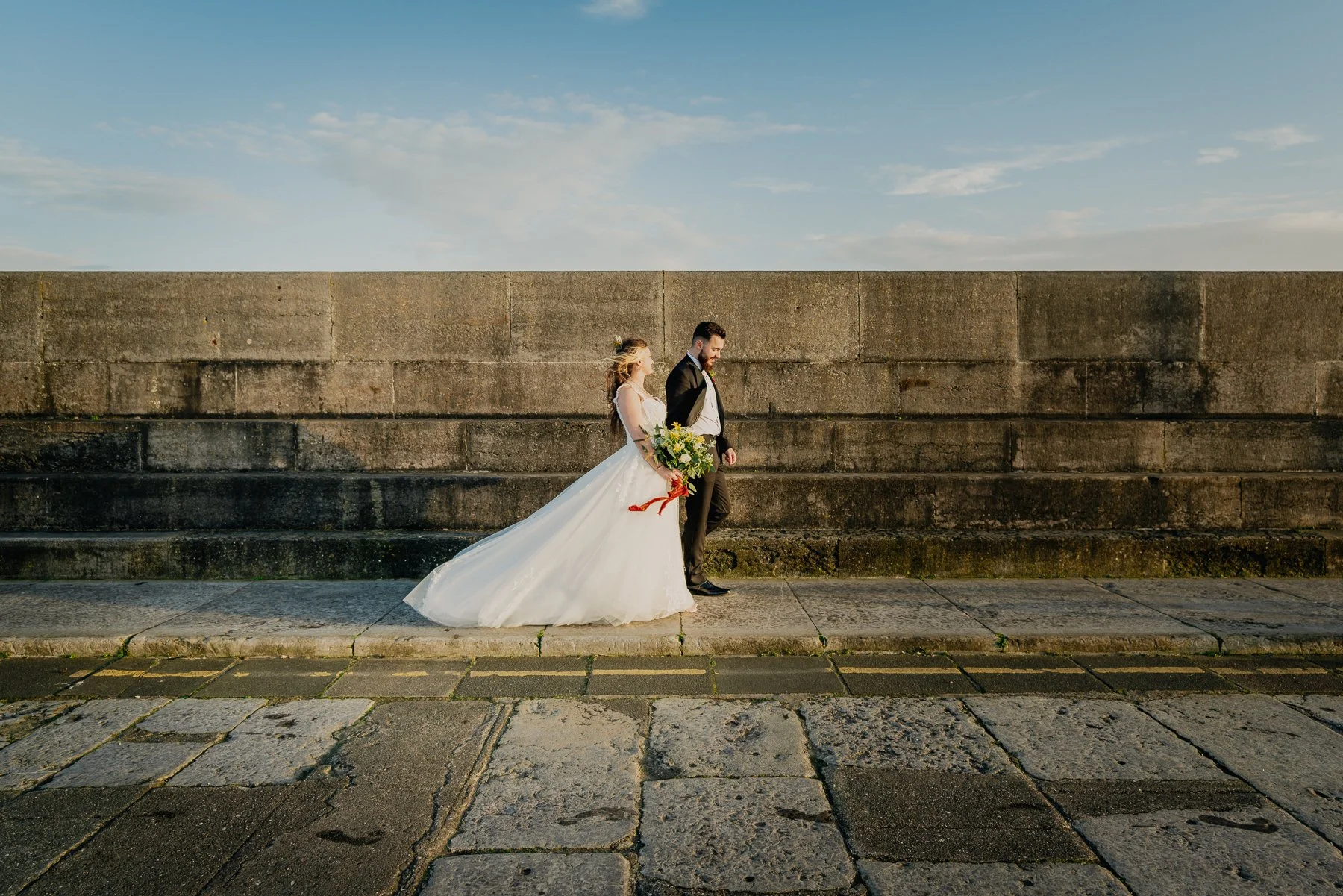 A bride and groom walking together along a stone sidewalk near a concrete wall, with the bride holding a bouquet of flowers, under a partly cloudy sky during sunset.