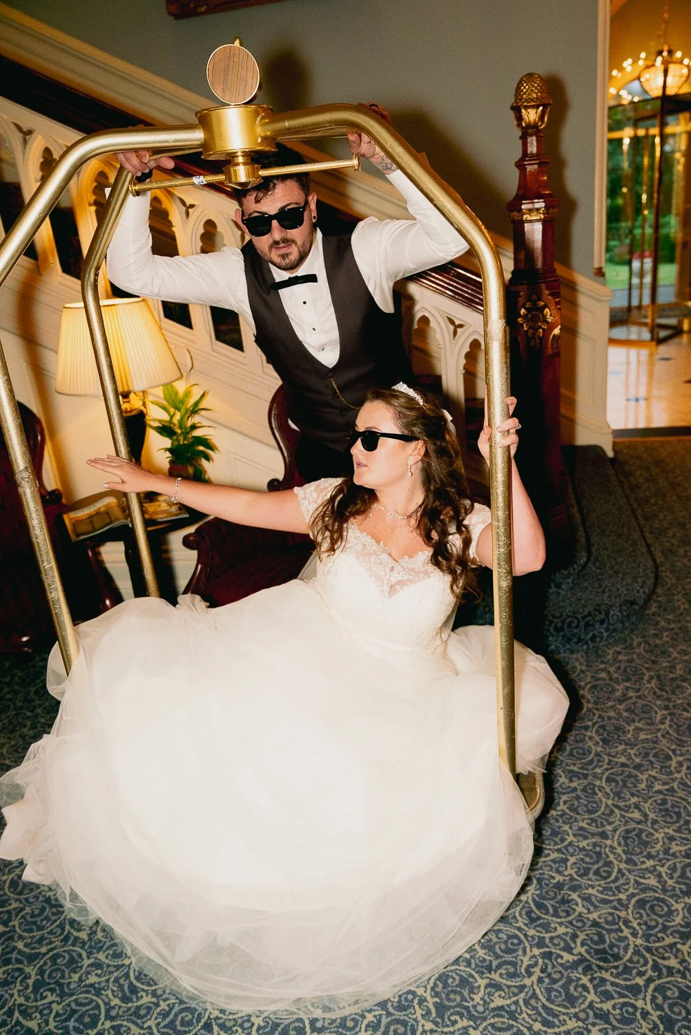 A bride in a white wedding dress and sunglasses sitting on a bench, and a groom in sunglasses, a vest, and bow tie standing behind her, both holding a gold luggage cart.
